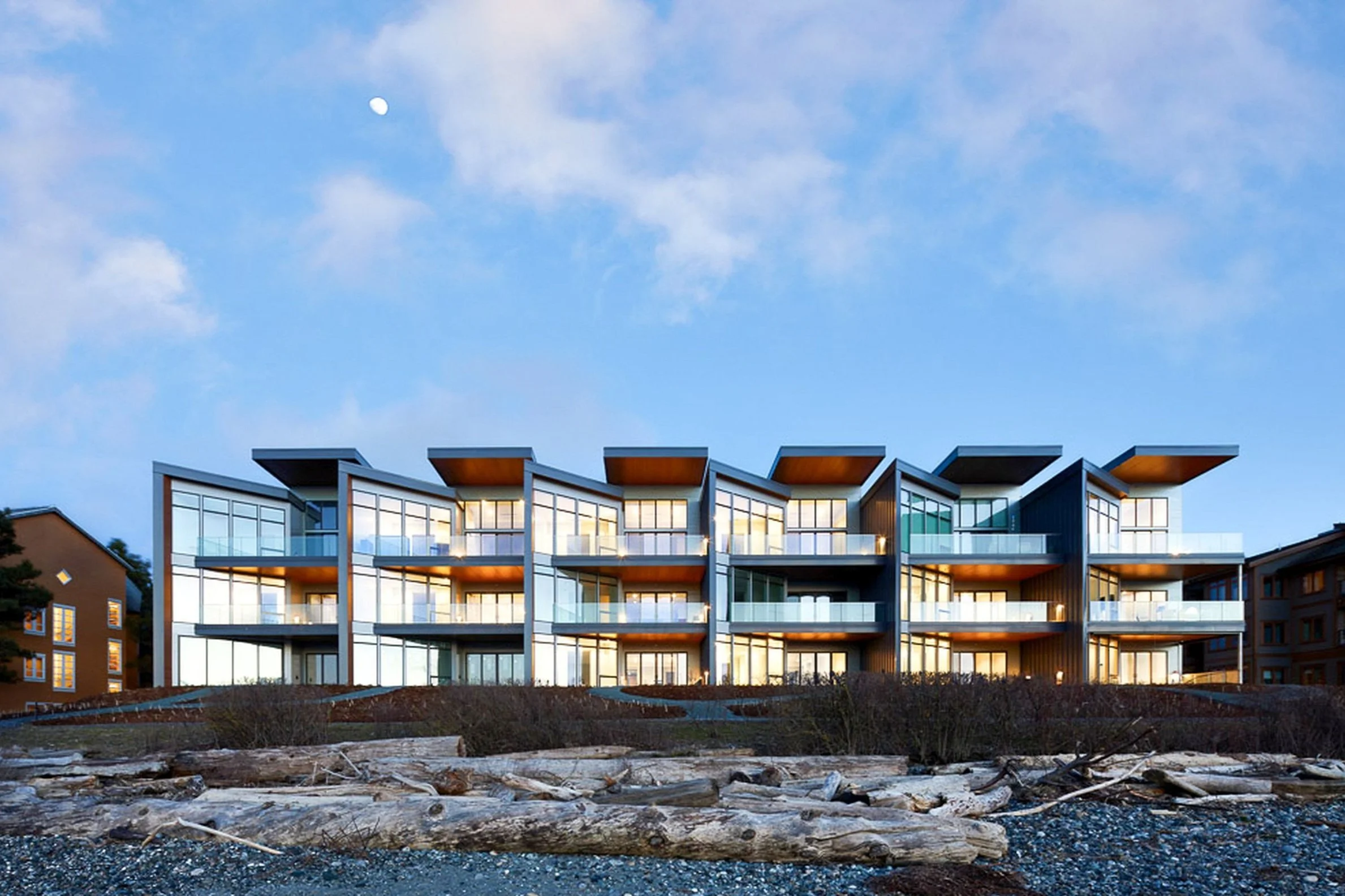 Modern multi-story apartment building with large glass windows and balconies, set against a blue sky with a visible moon.