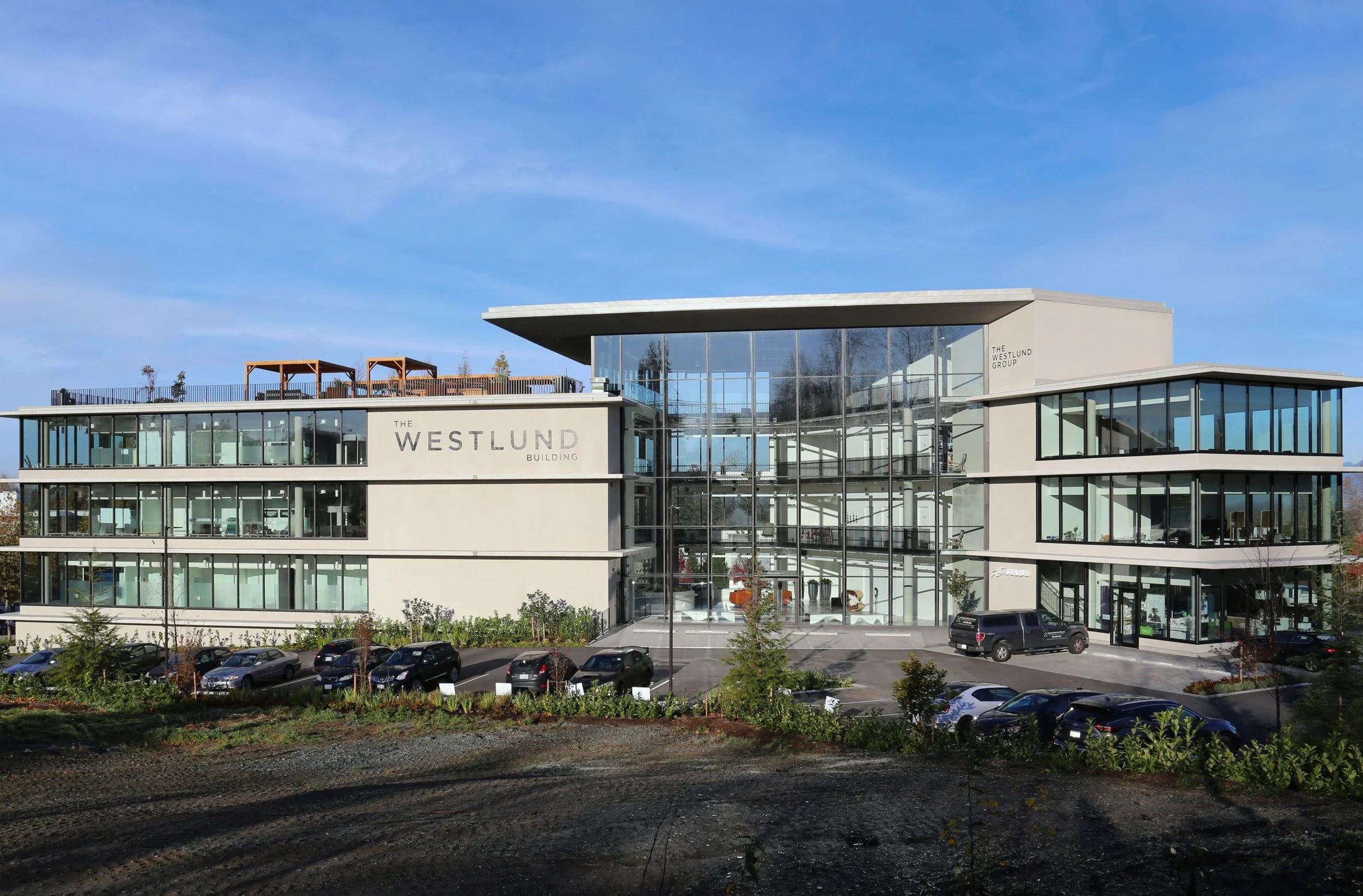 Modern multi-story office building with glass facade, labeled 'The Westlund Building,' and a parking lot with several cars in front.