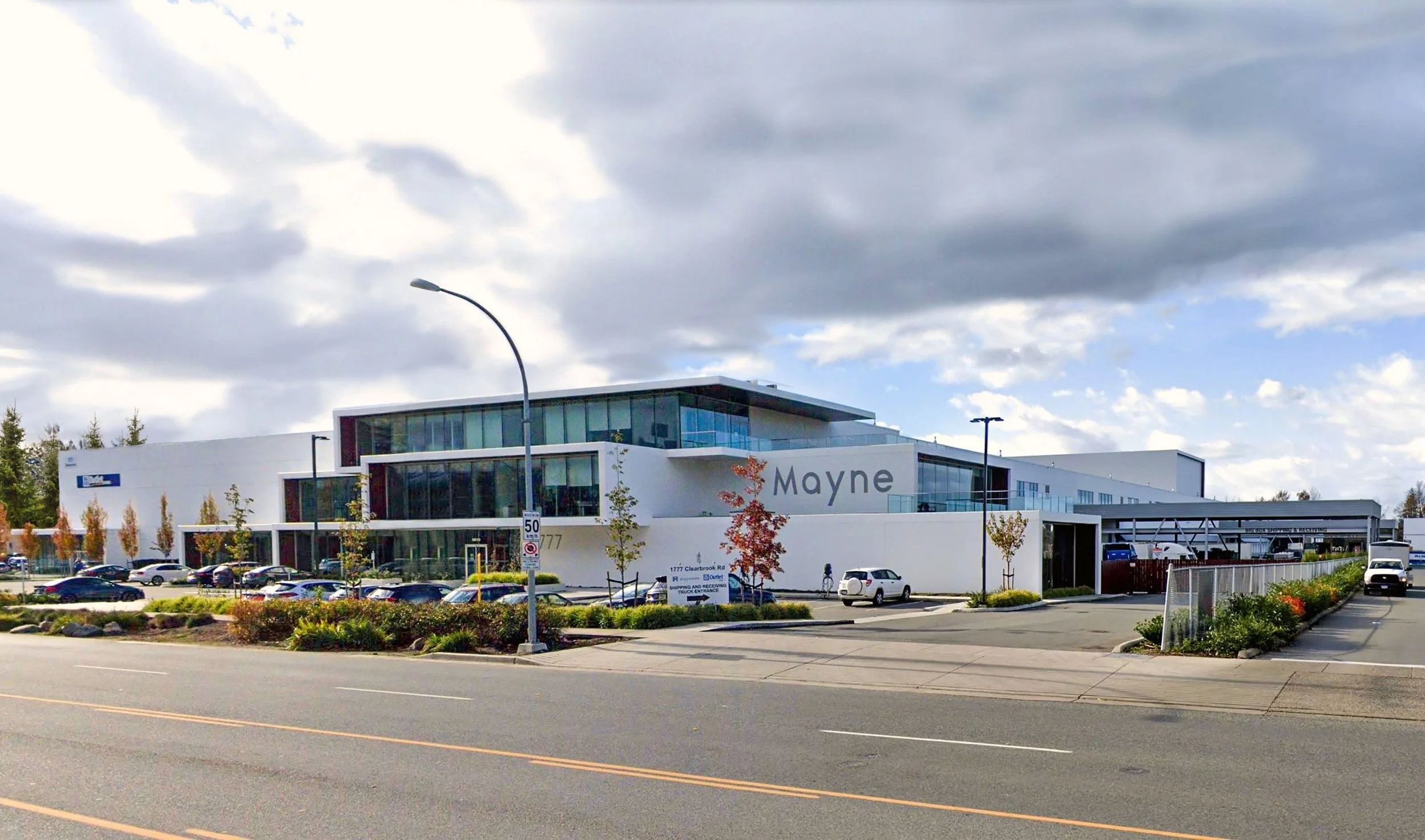 Modern commercial building with the name 'Mayne' on the side, parking lot in front, small trees, and cloudy sky overhead.
