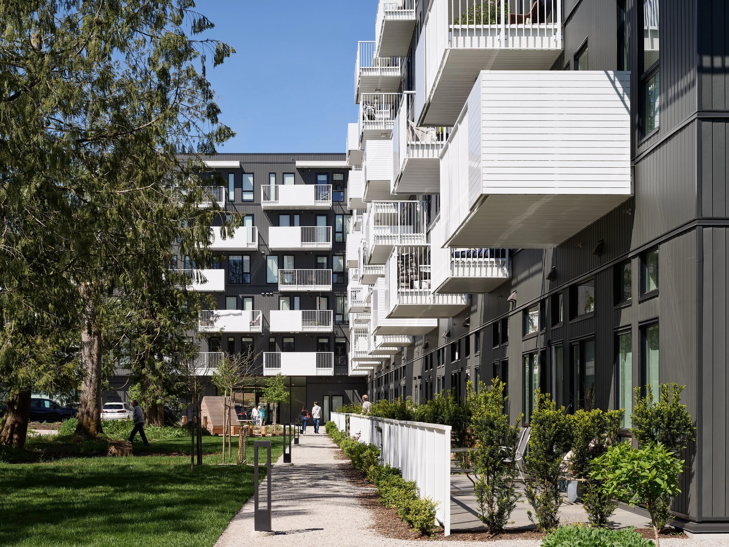 Modern multi-story apartment building with balconies, black and white exterior, surrounded by green trees and a landscaped walkway with people walking.