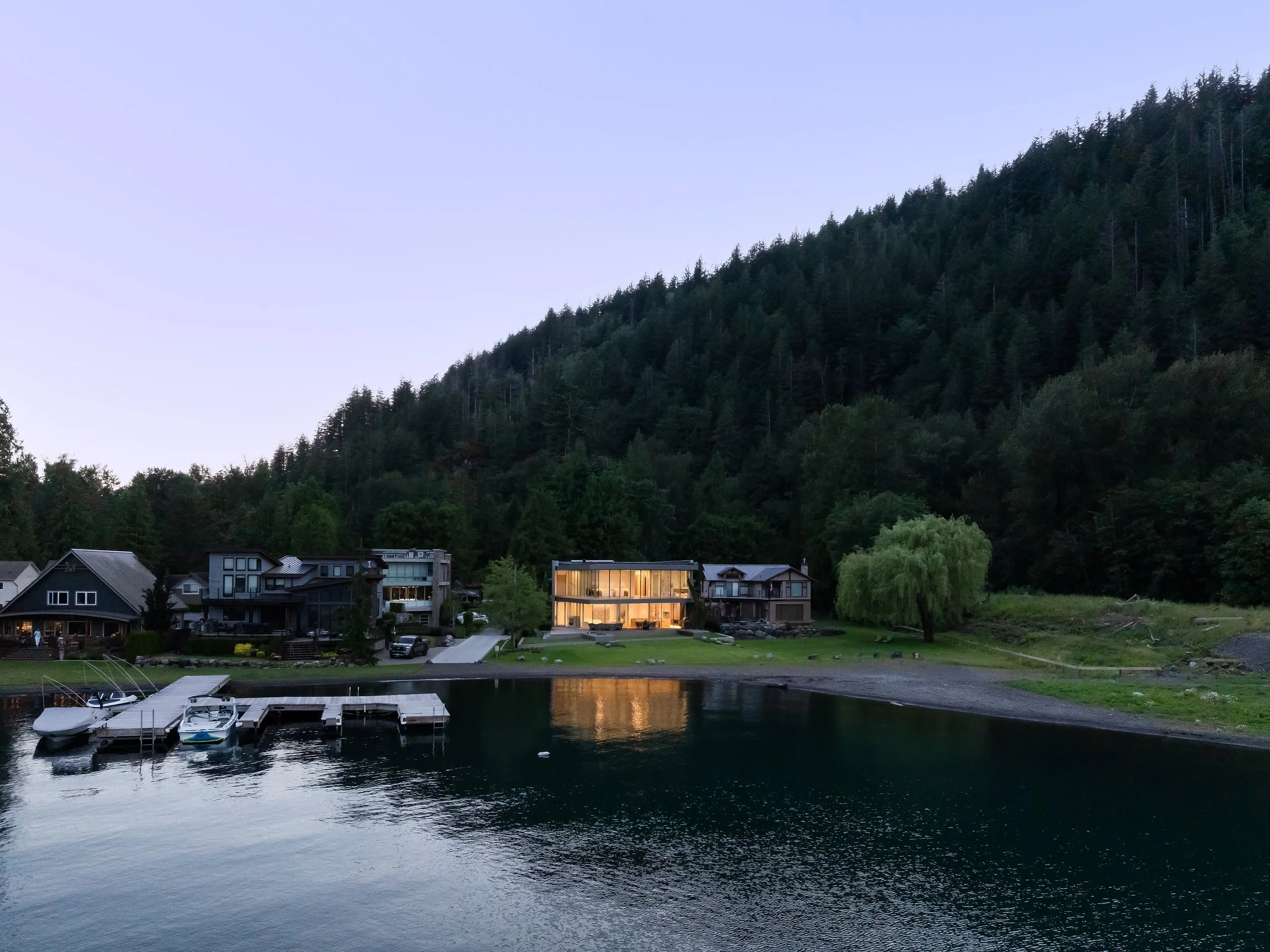 Residential houses on a lakeshore at dusk with a wooded hillside in the background and boats docked at a pier.
