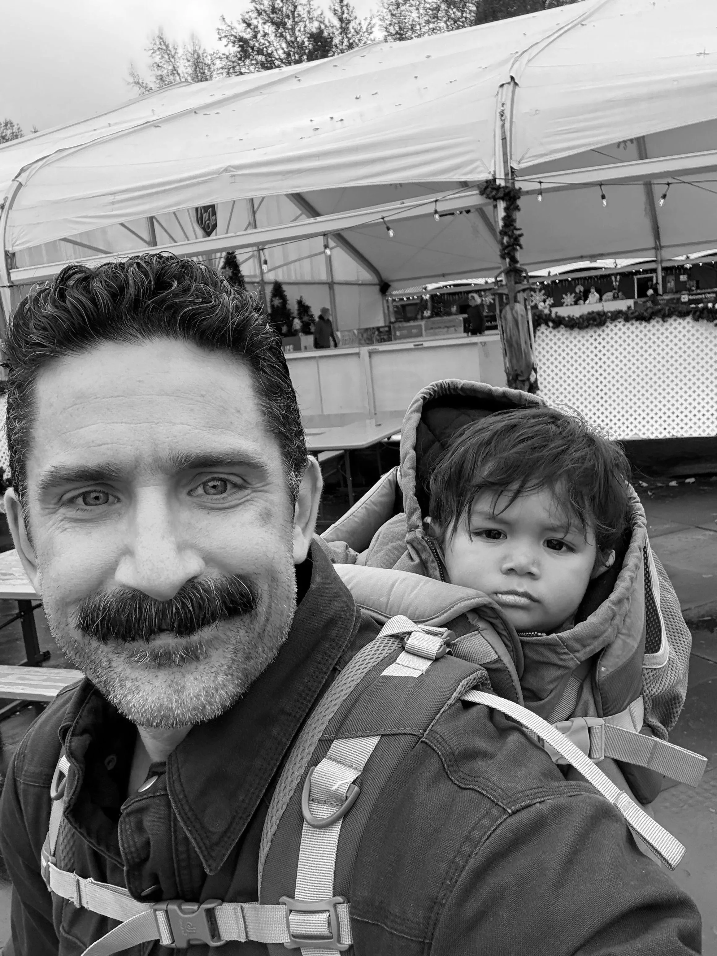 A man with a beard and a young child wearing a backpack at an outdoor ice skating rink.