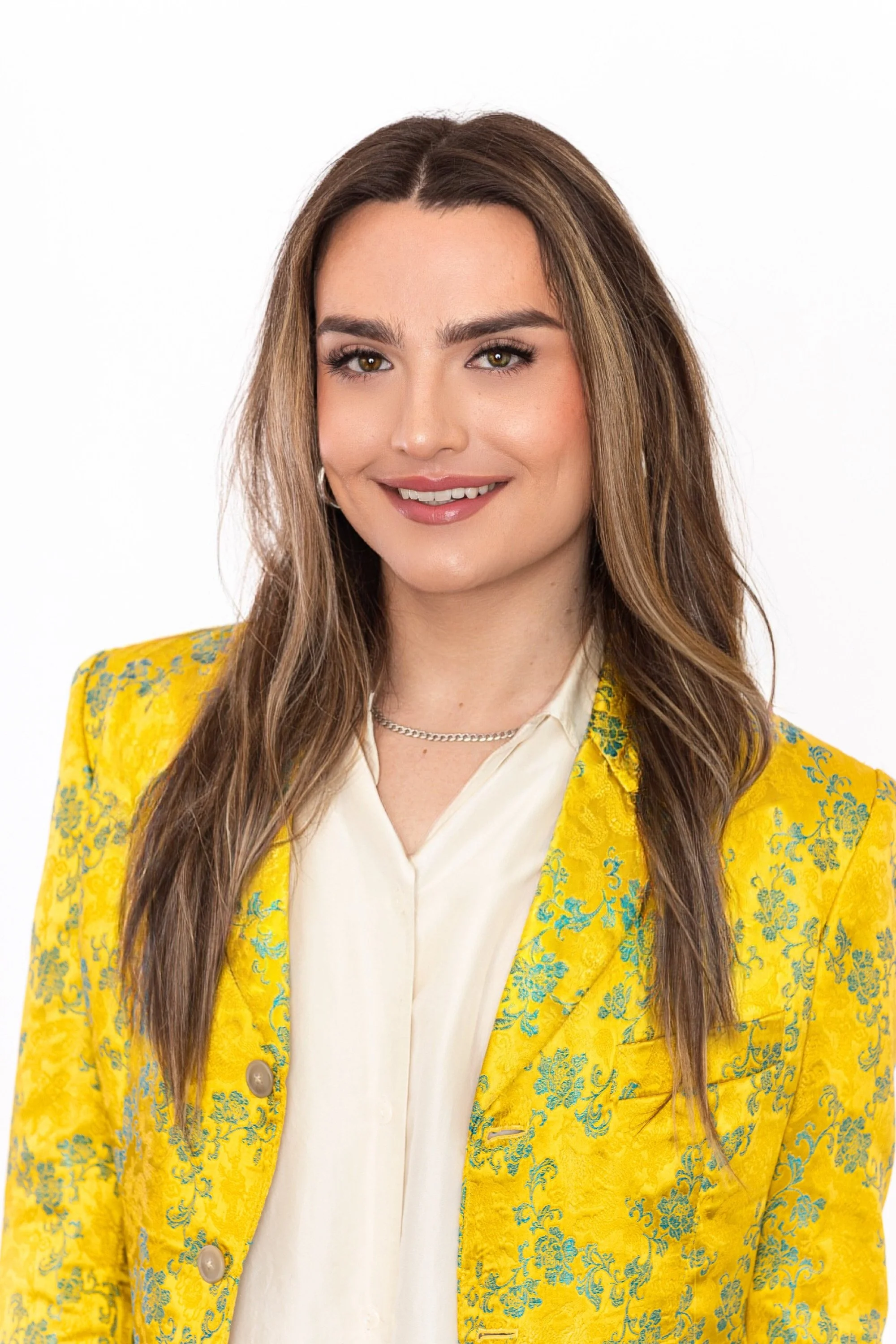 A woman with long brown hair and light makeup, smiling, wearing a yellow blazer with a floral pattern over a cream blouse, against a plain white background.