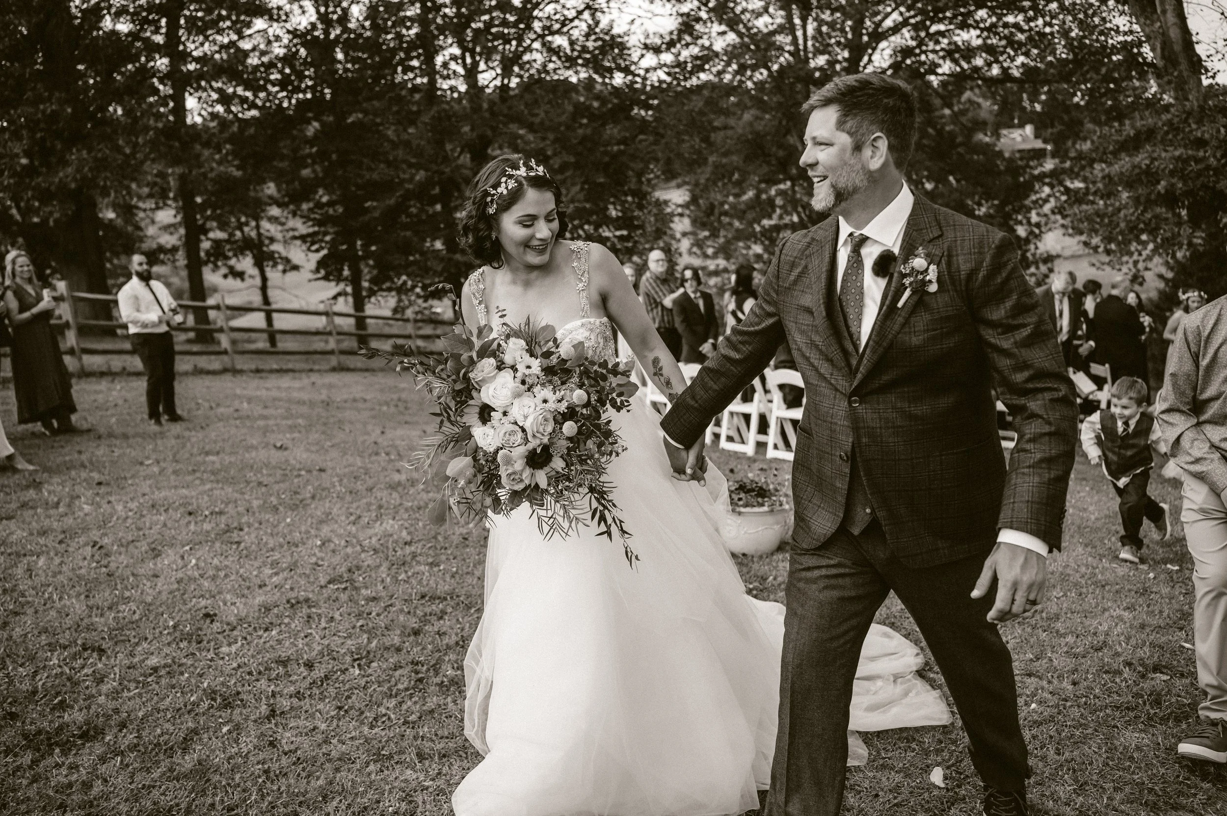 A bride and groom holding hands and smiling outdoors during their wedding, with guests in the background. Knoxville TN