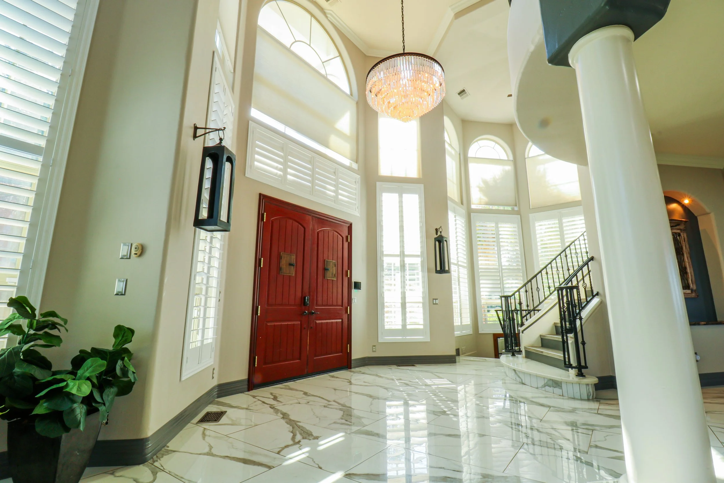 Luxurious front entrance with large marble tiled floor, double red wooden door, white shutters on windows, high ceiling with crystal chandelier, staircase with black railing, and large arched windows allowing natural light.