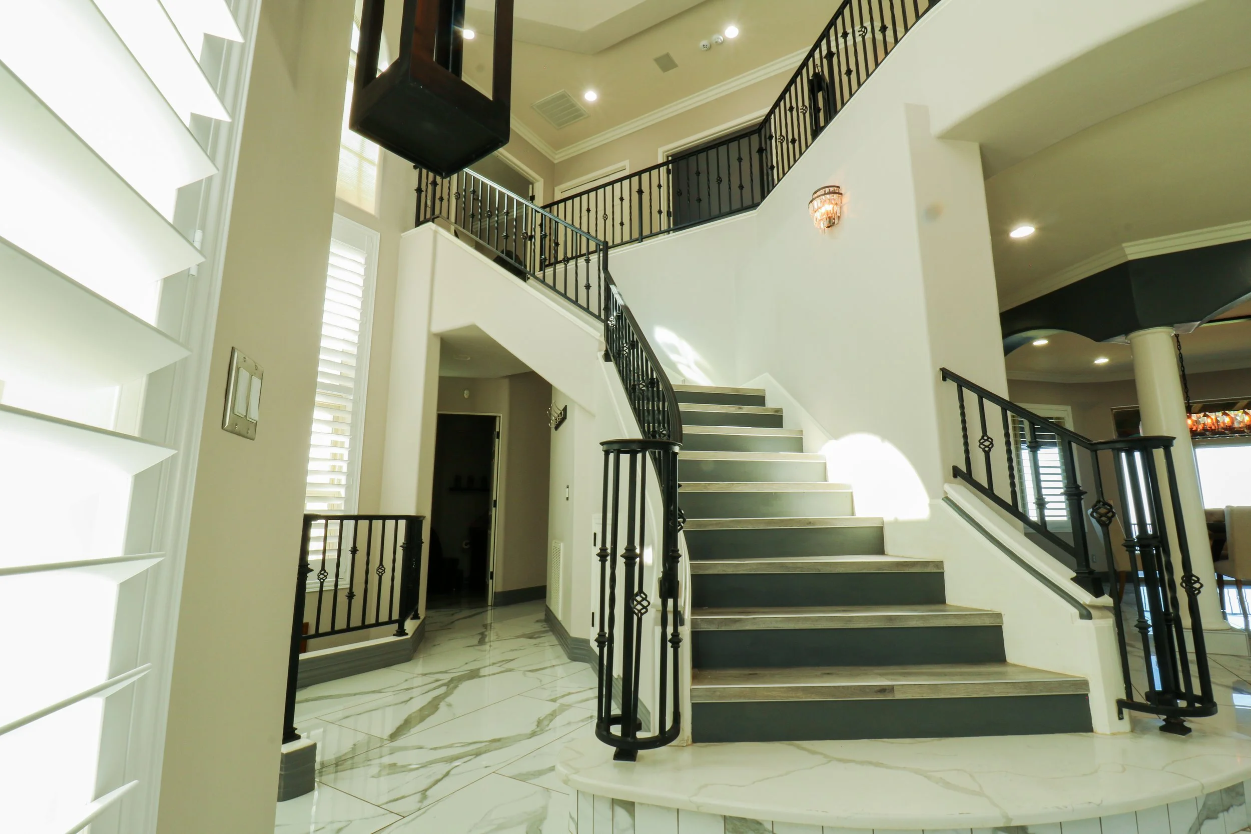 Interior view of a modern home featuring a marble floor, a staircase with black metal railings, and an open upstairs area with railing. Light fixtures and natural light are also visible.