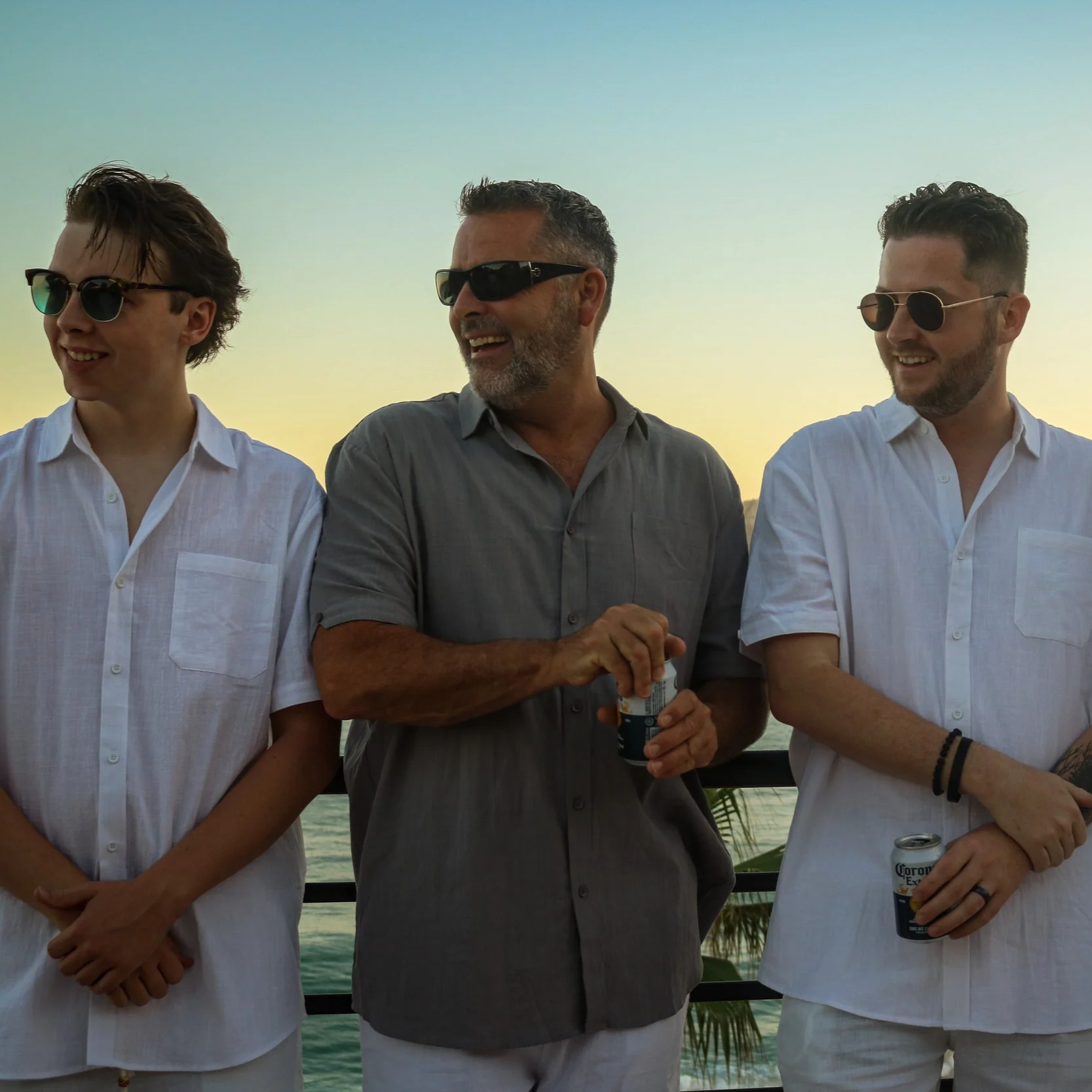 Four men standing by the water during sunset, talking and holding beer cans, wearing casual white and gray shirts, sunglasses, and smiling.