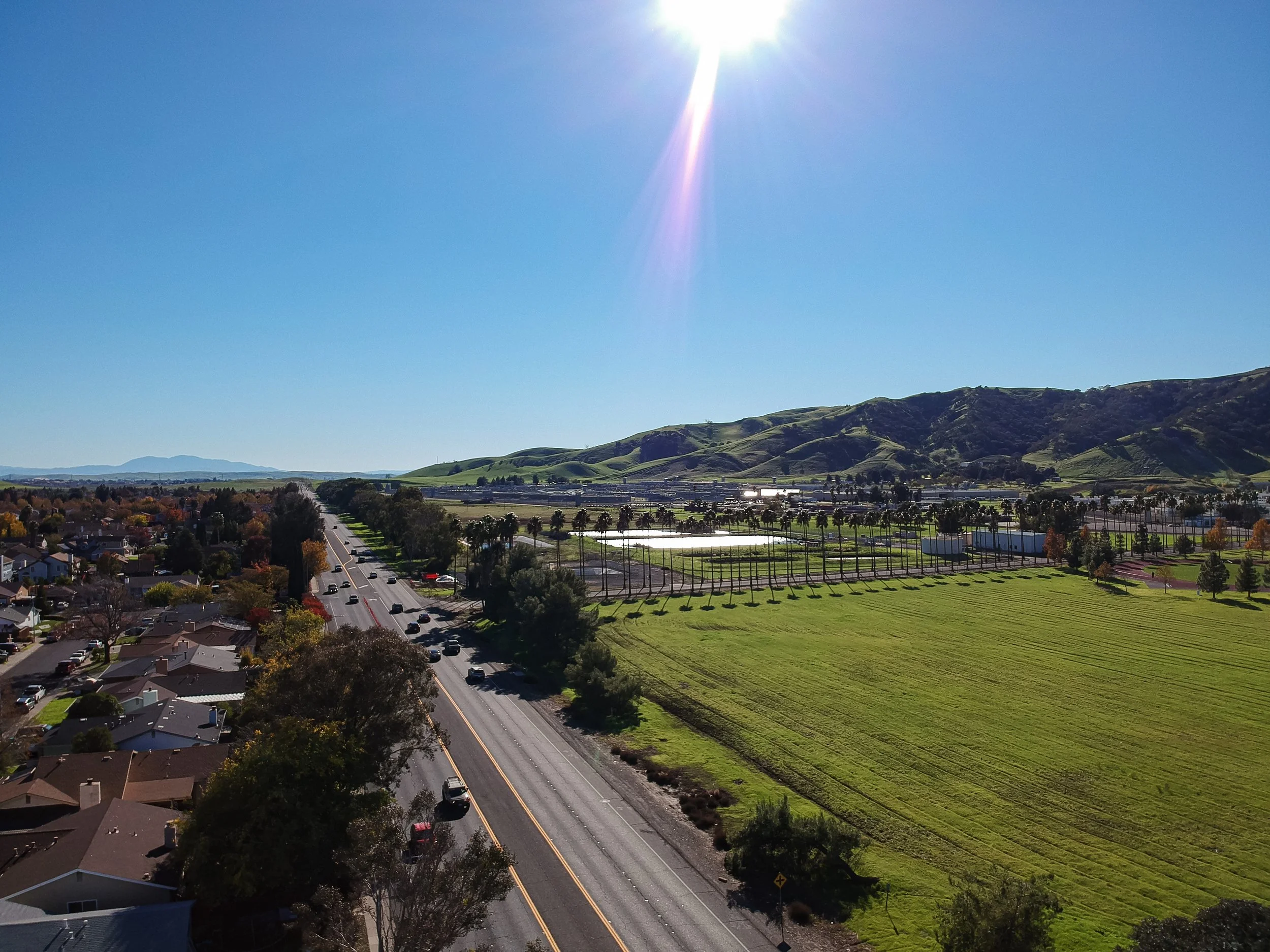 Aerial view of a suburban neighborhood with houses on the left, a two-lane road with cars, and open green fields with trees and a row of palm trees, with rolling green hills in the background and a bright sun in a clear blue sky.