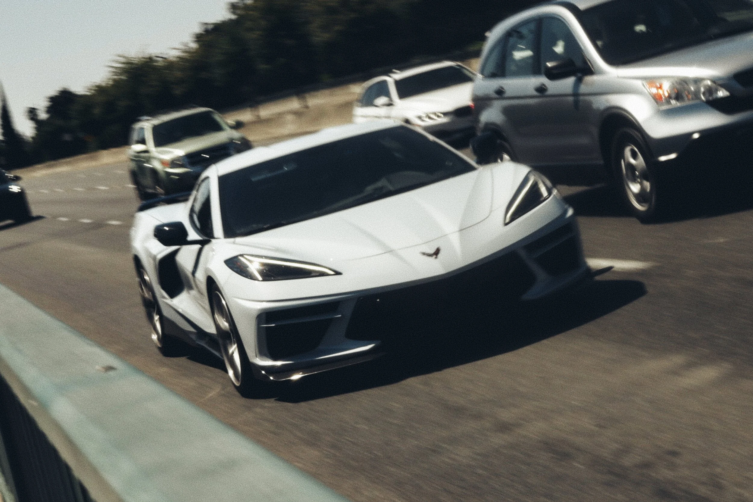 A white Corvette C8 driving on a multi-lane highway, surrounded by other vehicles including a Honda CRV.