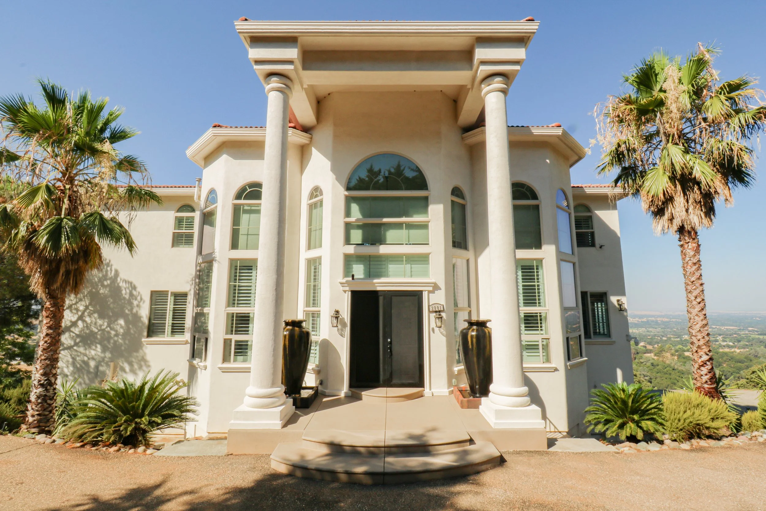 Front view of a large, modern, white house with tall columns, arched windows, and decorative planters, surrounded by palm trees under a clear blue sky.