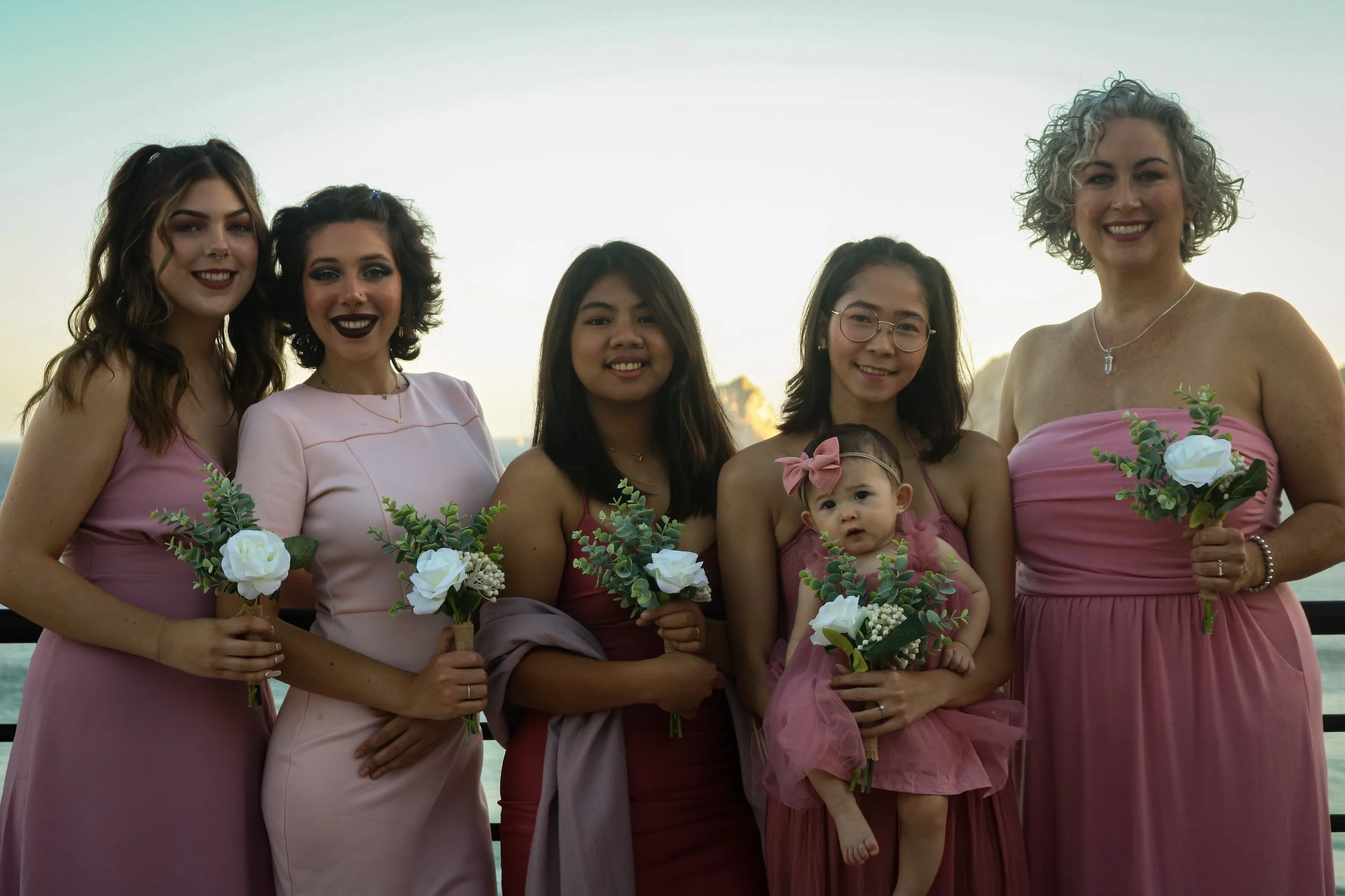 Group of six women and a young girl standing outdoors in evening light, each holding a small bouquet of white flowers and greenery, dressed in pink dresses, with a body of water and sky in the background.