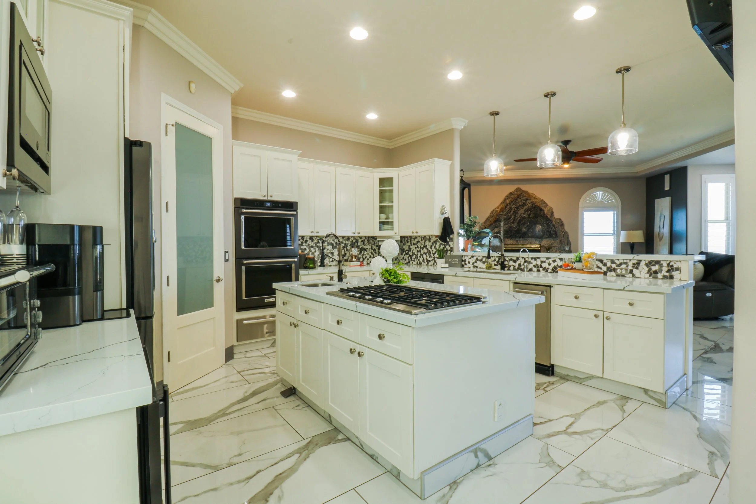 Modern kitchen with white cabinets, marble floors, and a central island with a gas cooktop, overlooking a living room with a rock feature wall and ceiling fan.