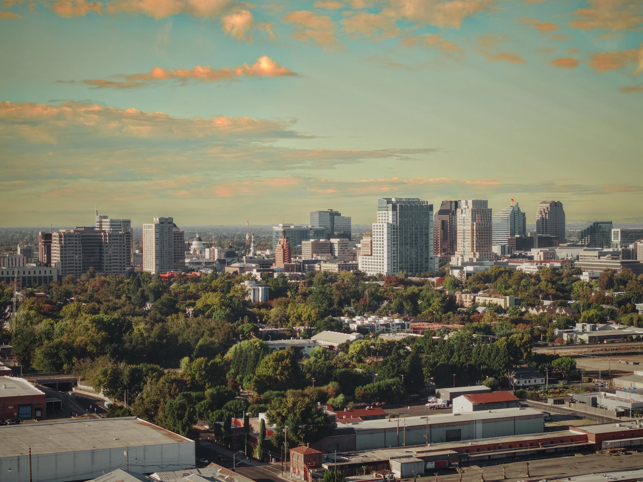 Sacramento, CA skyline with various skyscrapers and buildings surrounded by green trees, under a partly cloudy sky at sunset.