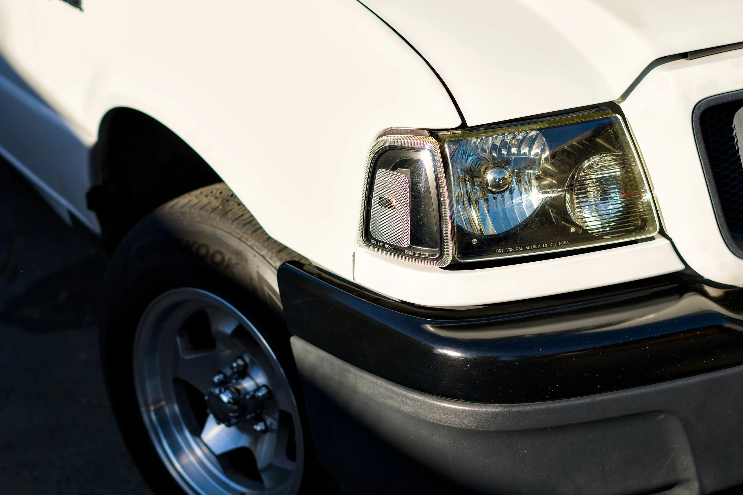 Close-up of the front right corner of a white Ford Ranger, showing the headlight, part of the black bumper, and a silver wheel with a tire.