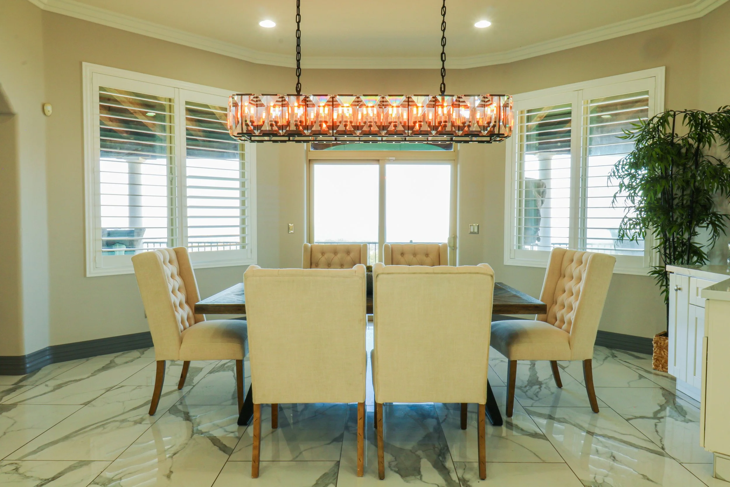 Dining room with a large wooden table surrounded by eight beige upholstered chairs, a modern rectangular chandelier hanging above, marble tiled floor, and windows with white plantation shutters. There is a sliding glass door in the background and a t