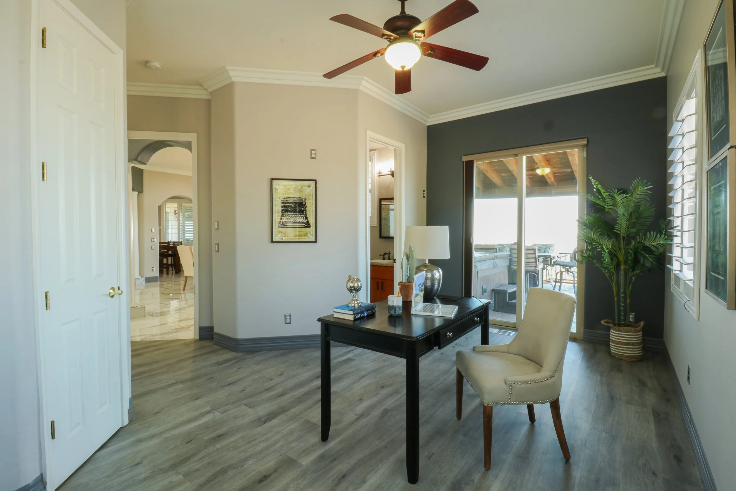 Interior view of a living room with light-colored walls, a dark accent wall, gray wood flooring, a ceiling fan with wooden blades, a black desk with a beige chair, a table lamp, decorative items, potted plant, and sliding glass door leading to an out