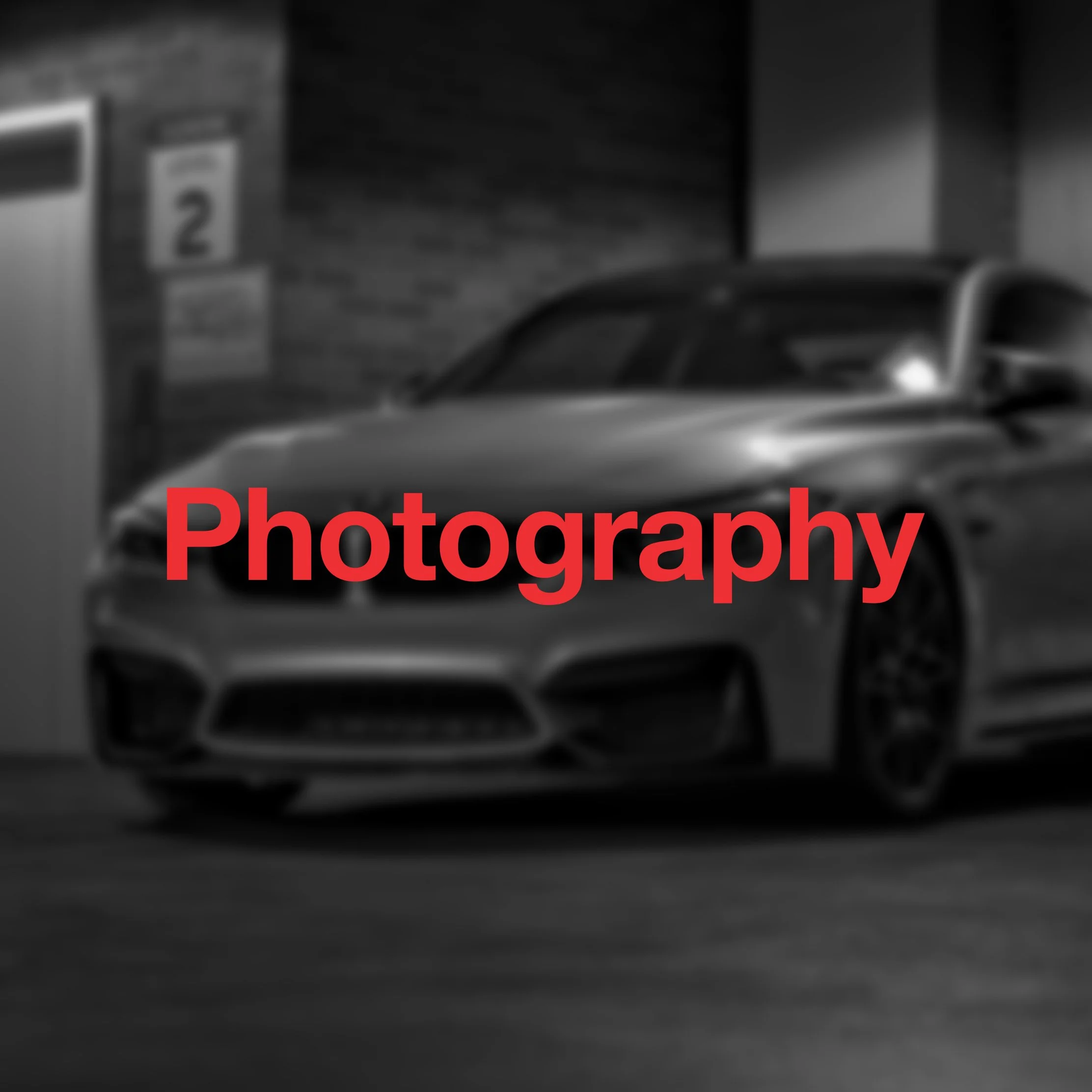Black and white photo of a sports car parked next to a brick building with a sign that says '2' and some other text. The word 'Photography' is written in red over the image.
