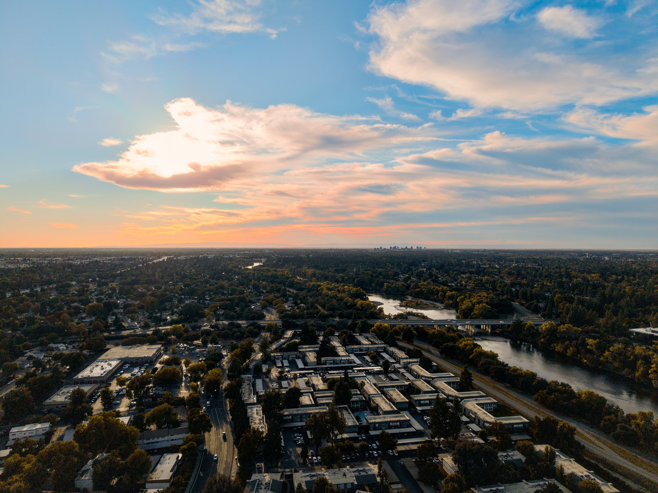 Aerial view of Sacramento in the distance with a river, bridges, trees, and buildings at sunset with a partly cloudy sky.