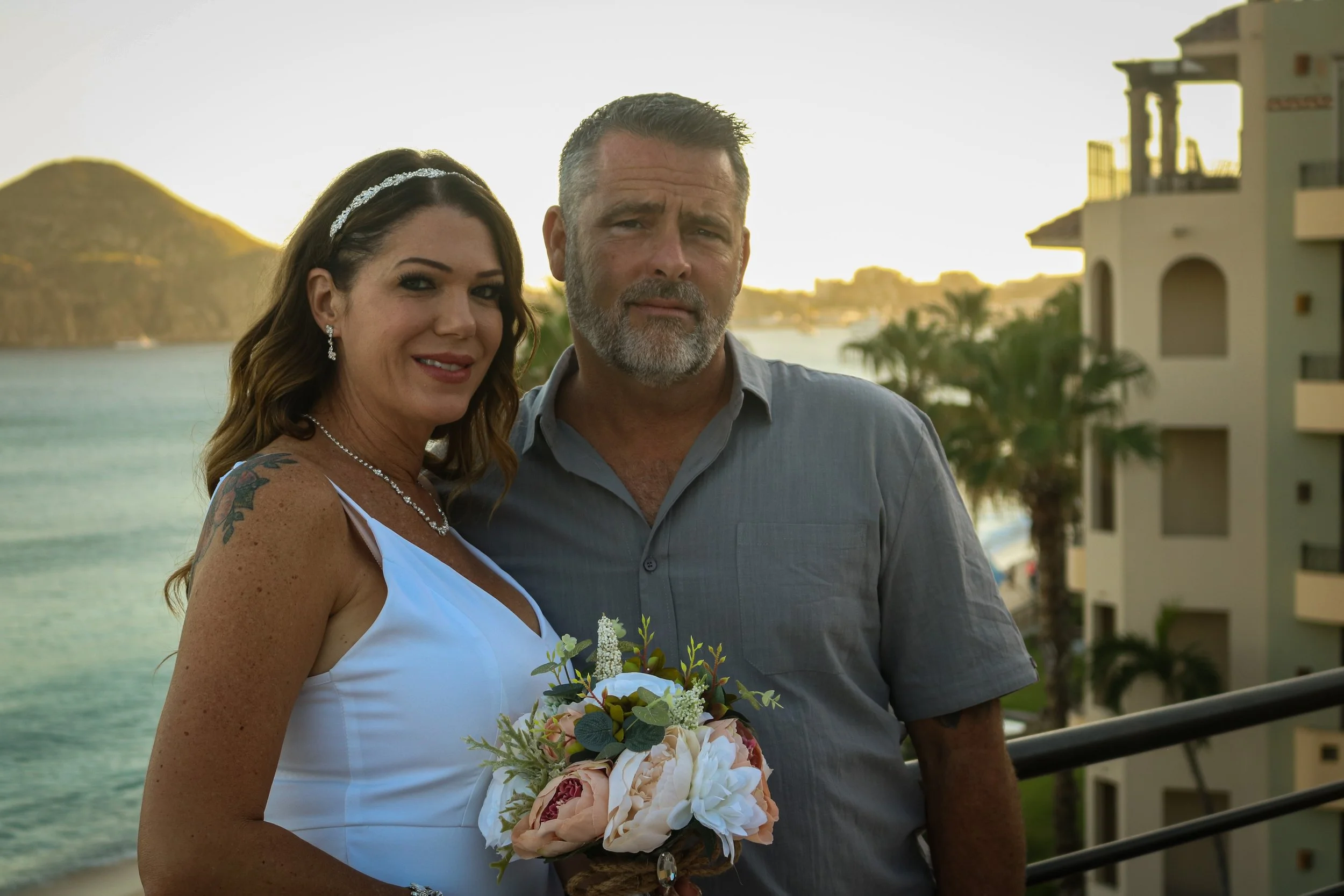 A middle-aged woman in a white dress holding a bouquet of flowers, standing next to a man in a gray shirt, at a beachside location with water, palm trees, and buildings in the background during sunset.