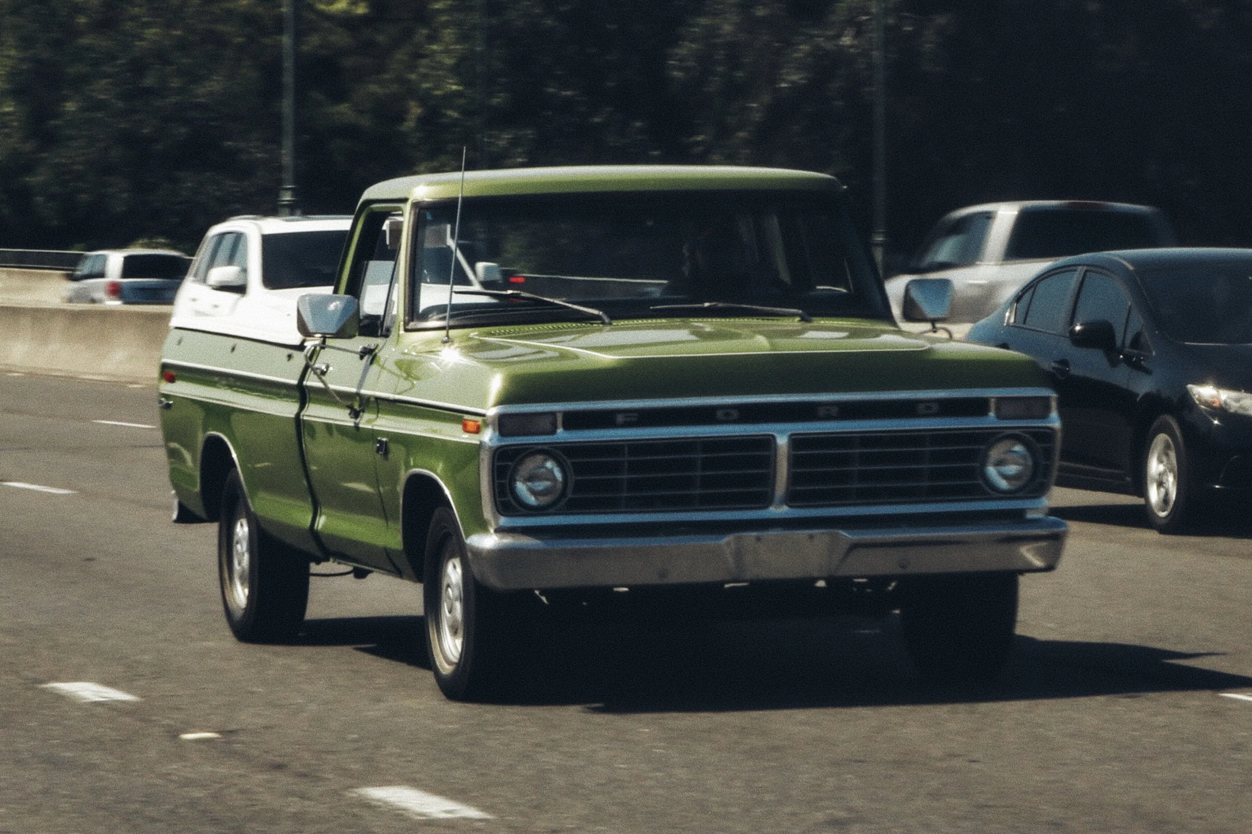 A vintage green and white Ford pickup truck driving on a highway with other cars in the background.