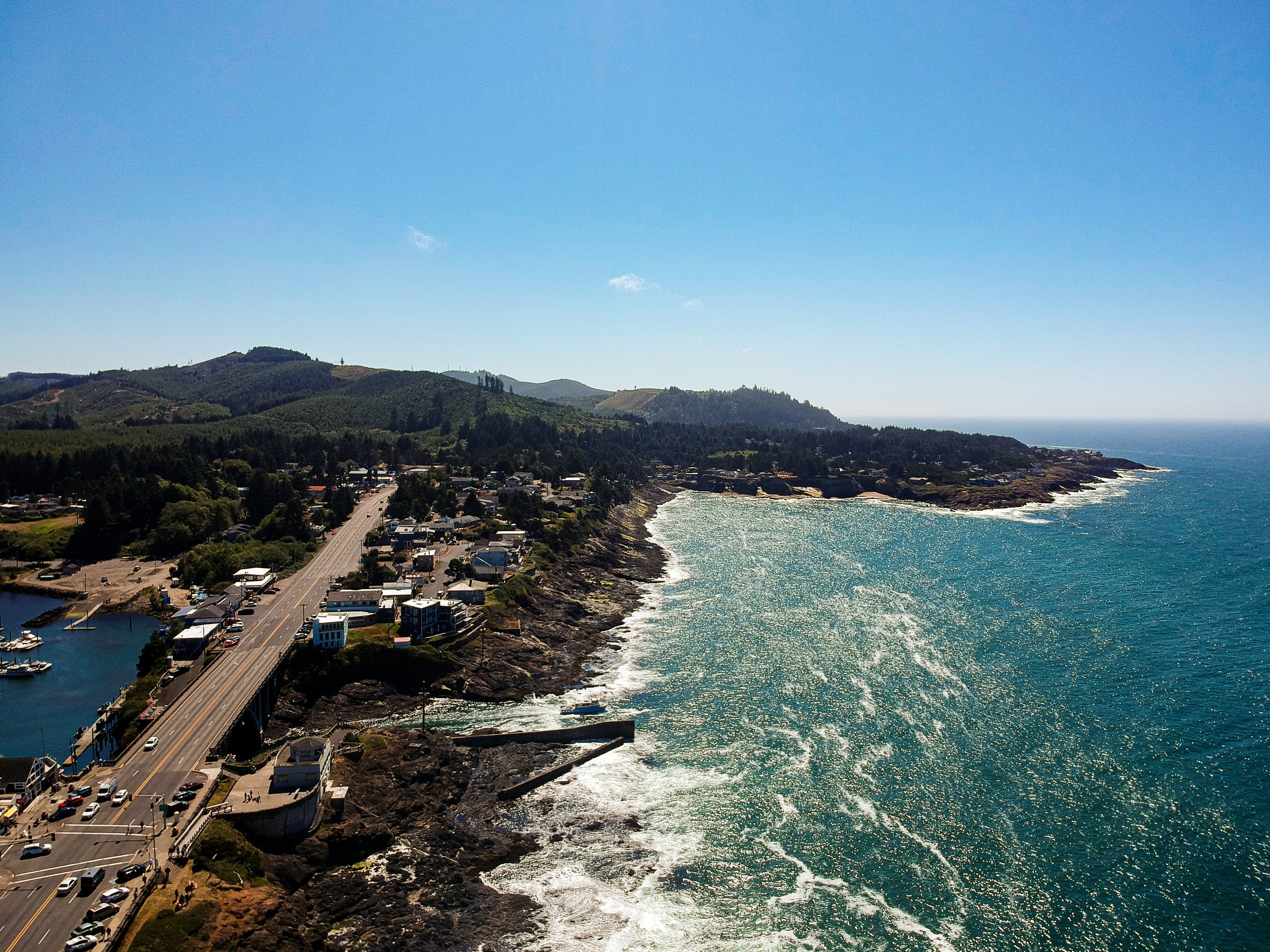 Aerial view of a coastal town with a bridge over the water, surrounded by green hills and the ocean under a clear blue sky.