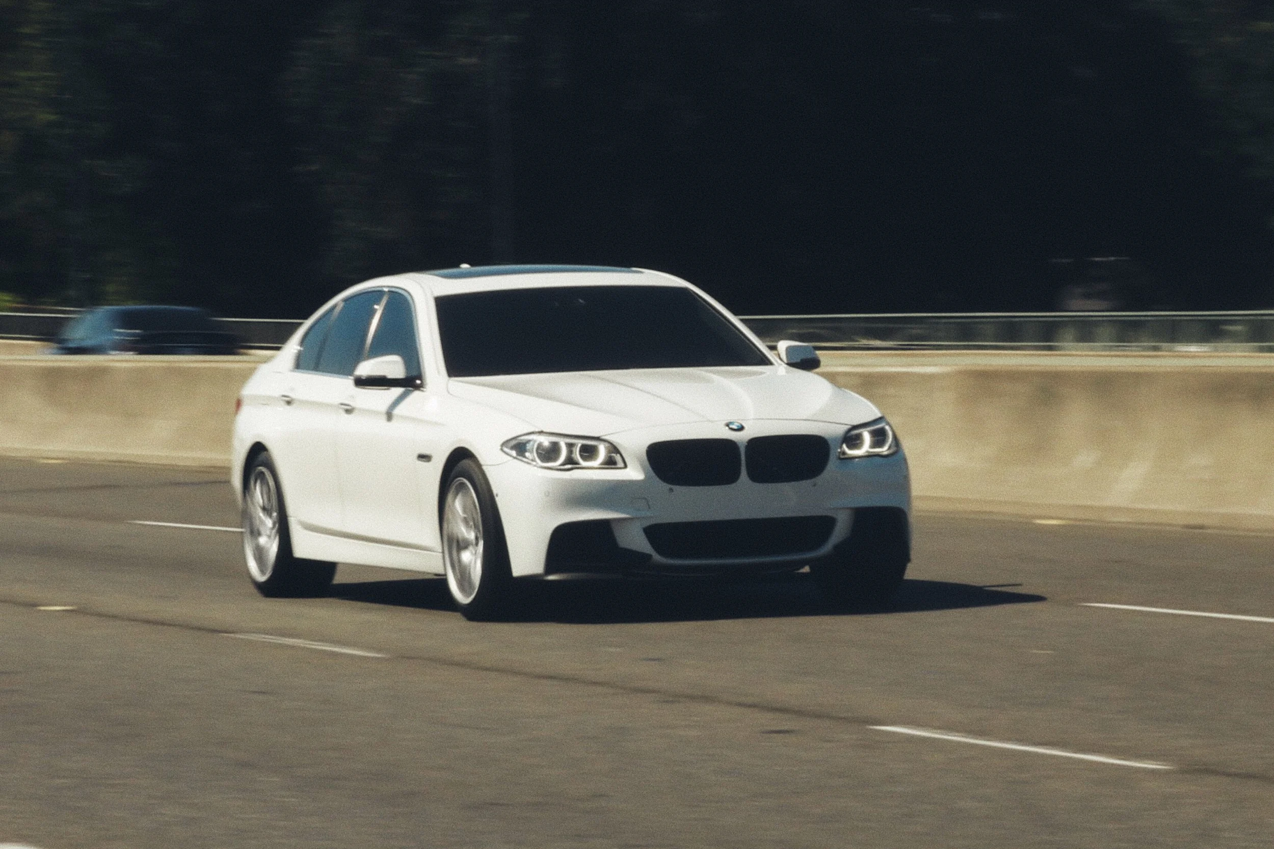 White BMW 535i M-Sport sedan driving on a highway with a concrete barrier and trees in the background.
