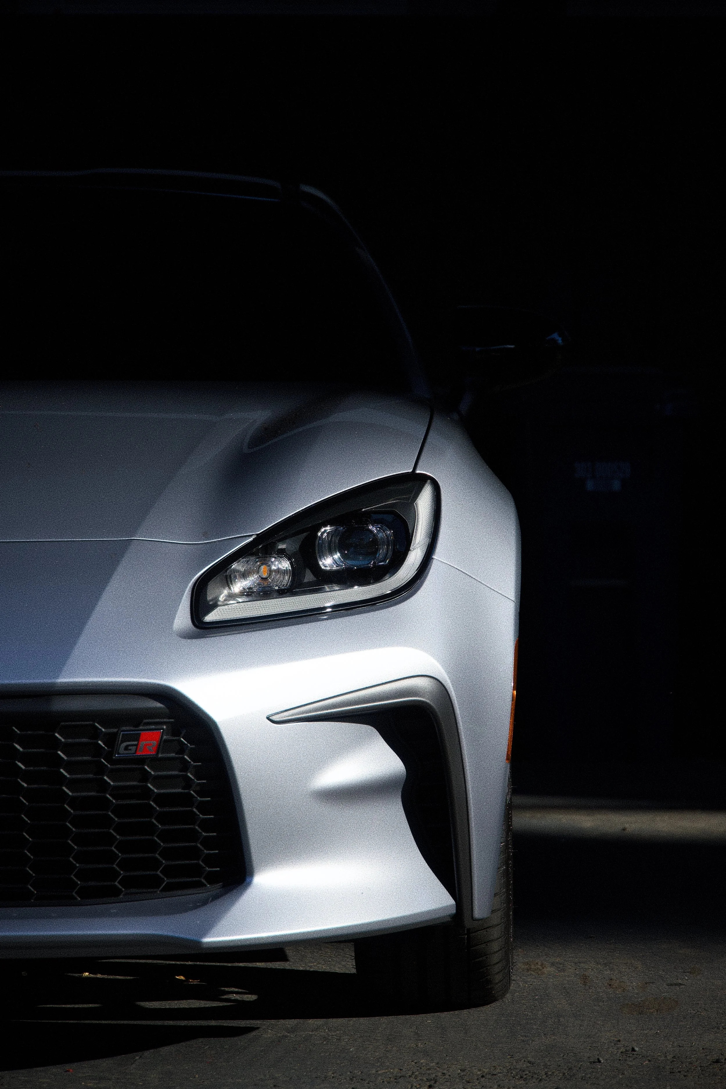 Front-right view of a silver sports car in a dark setting, showing the headlight, part of the front grille with a red 'GR' badge, and the tire.