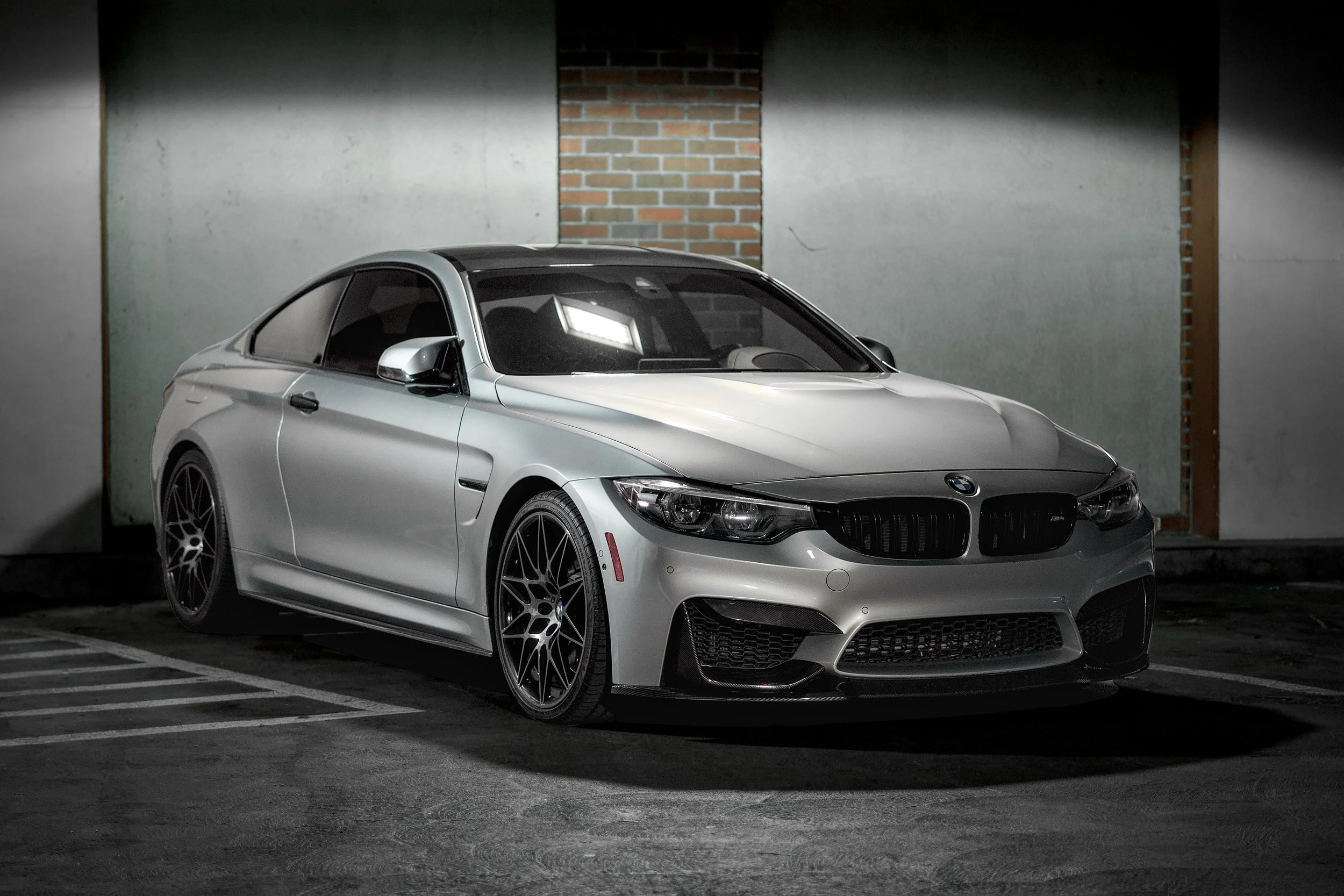 Silver BMW sports car parked in a dimly lit parking lot at night, with a brick wall and concrete wall background.