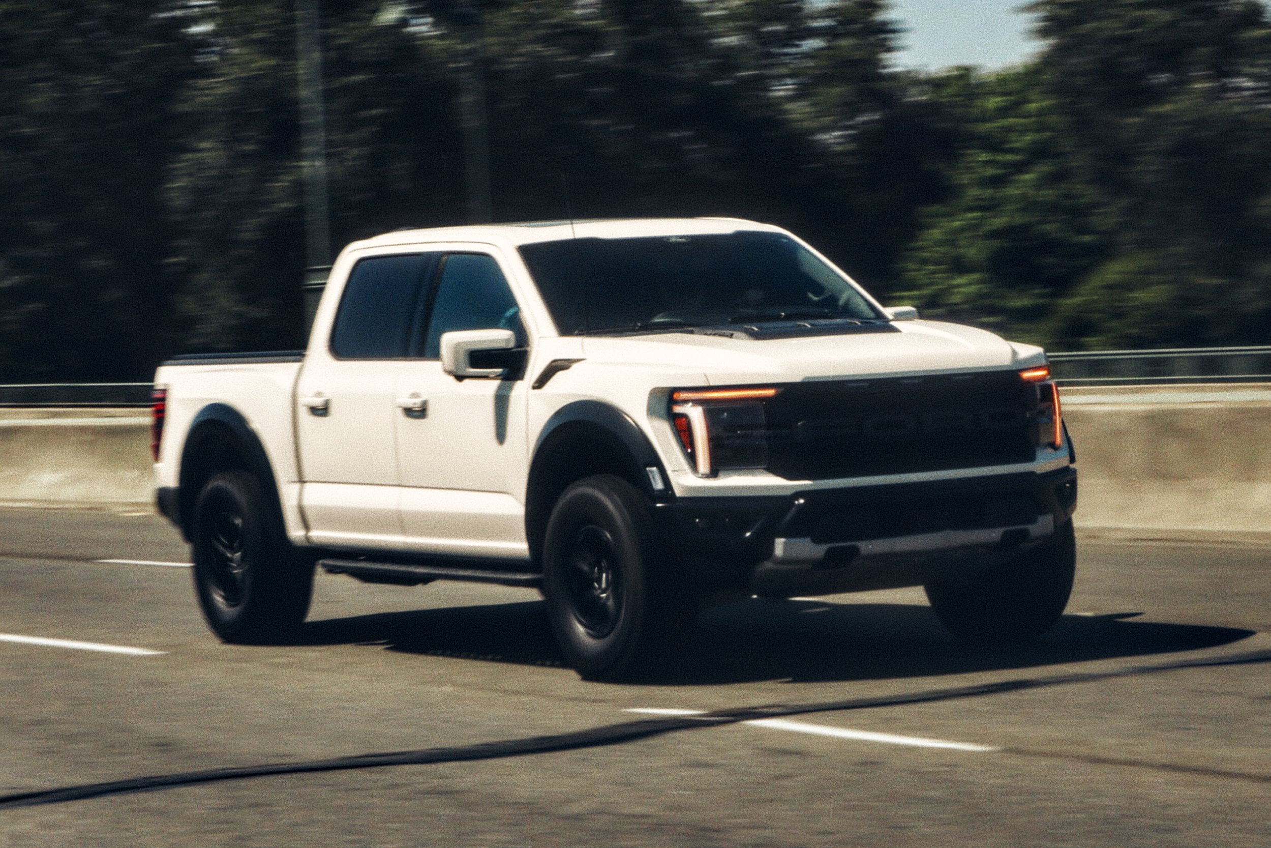 A white Ford F-150 Raptor driving on a highway with trees in the background.