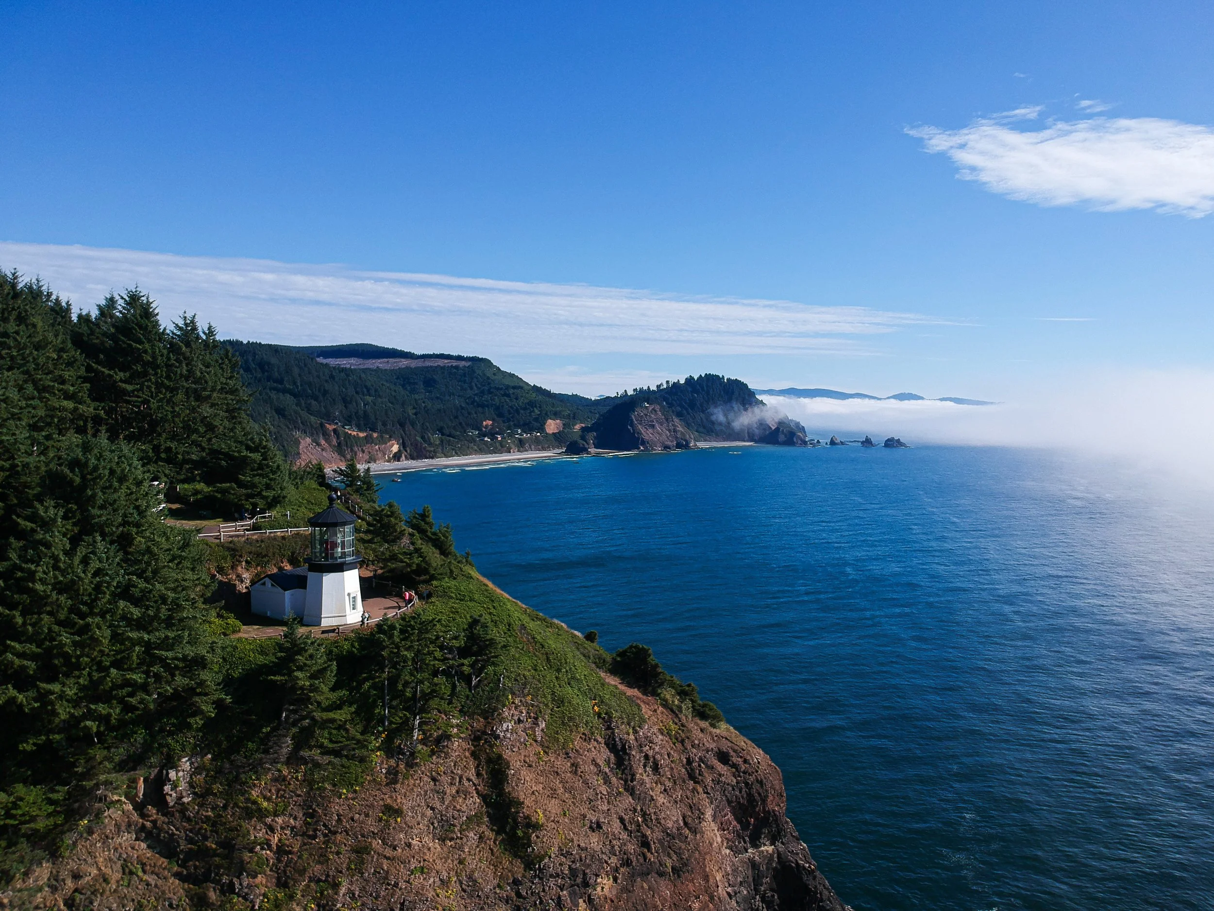 Coastal landscape with a lighthouse on a grassy hill in Netarts, OR, surrounded by trees, overlooking the Pacific ocean with cliffs and distant islands under a partly cloudy sky.