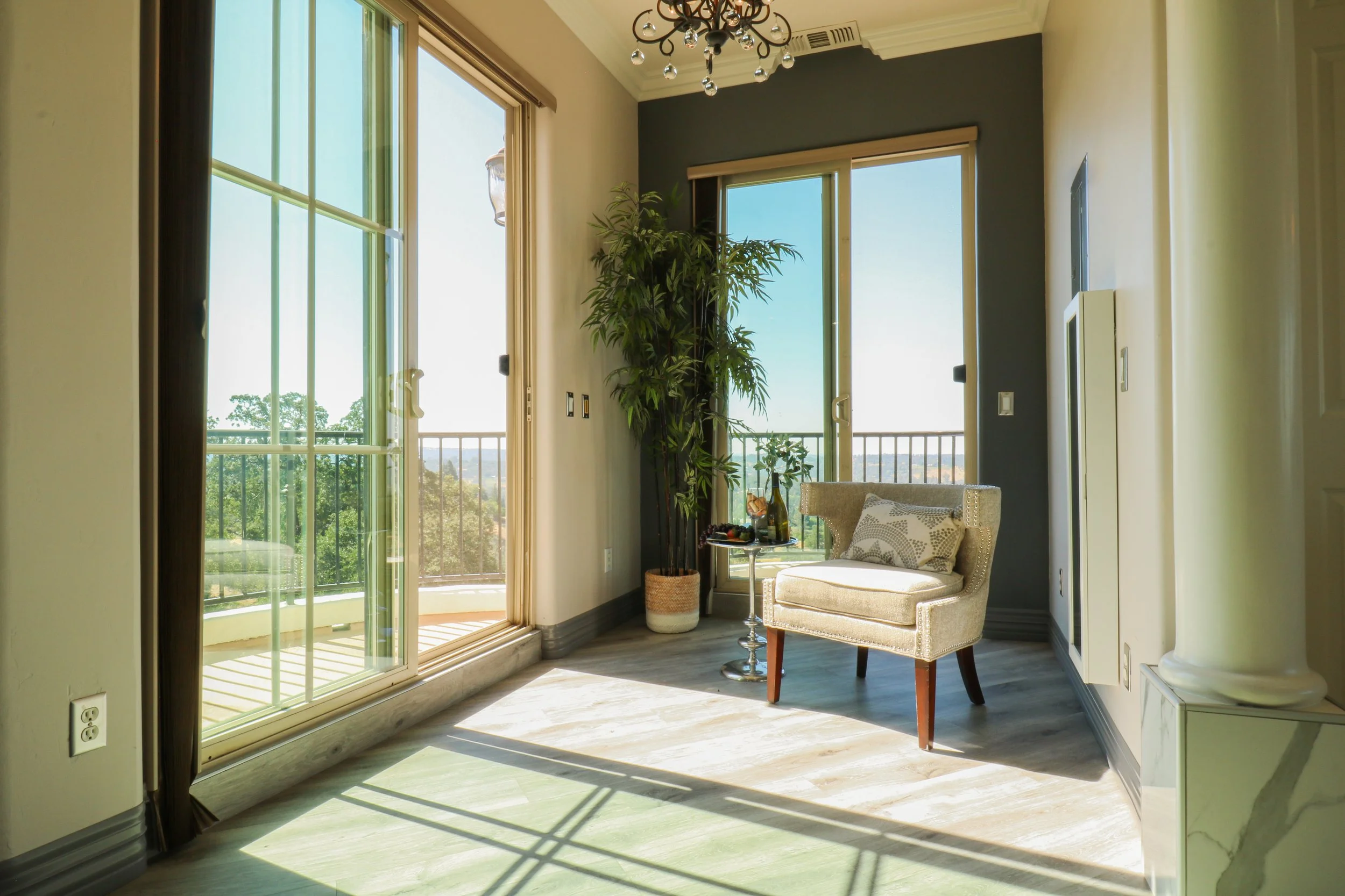 Bright living room with large sliding glass doors leading to a balcony, a beige armchair with pillows, a potted plant, sunlight casting shadows on the wooden floor.