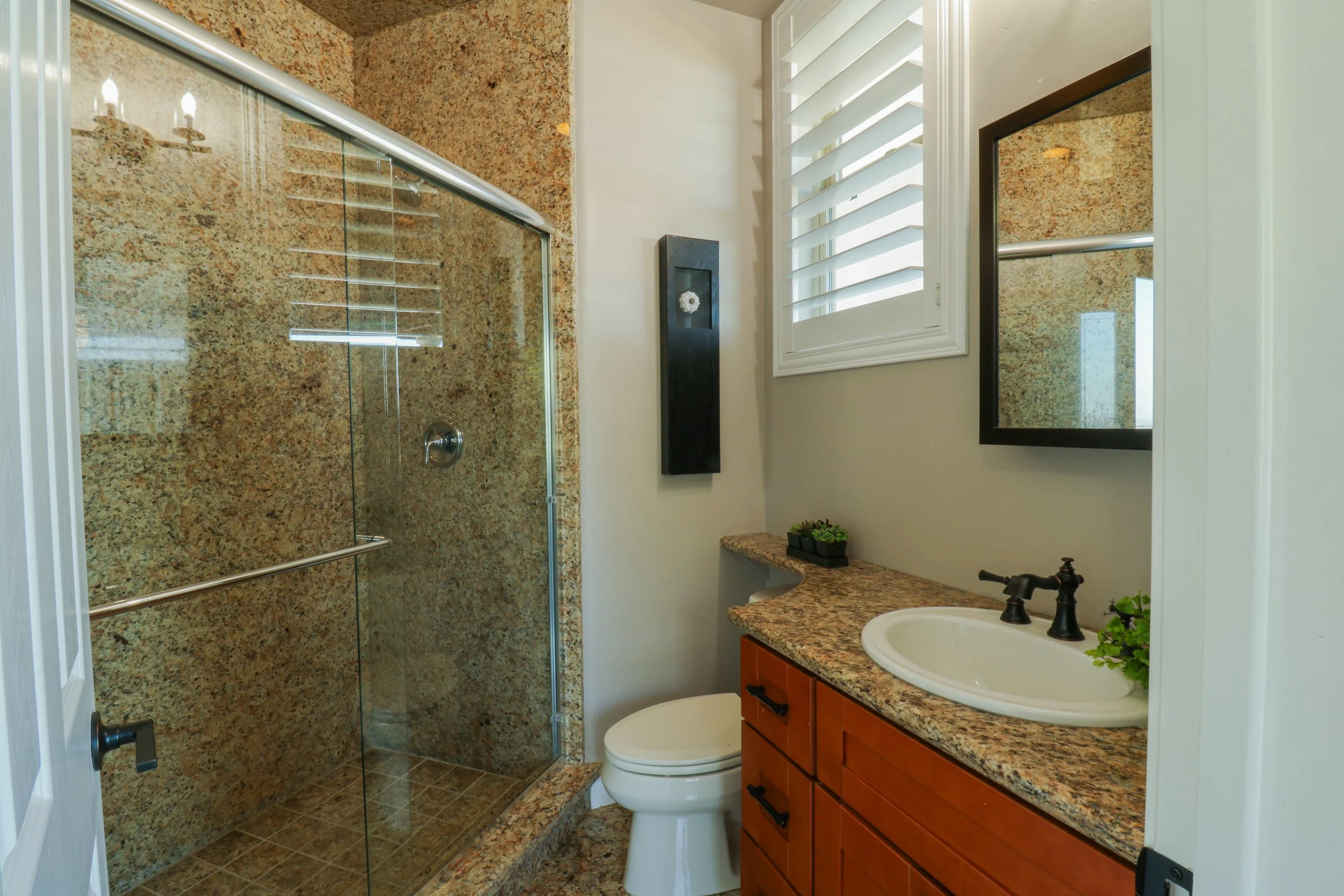 Bathroom with a glass shower enclosure, a granite countertop with a sink, a mirror, a window with shutters, a small plant, and a black wall decoration.