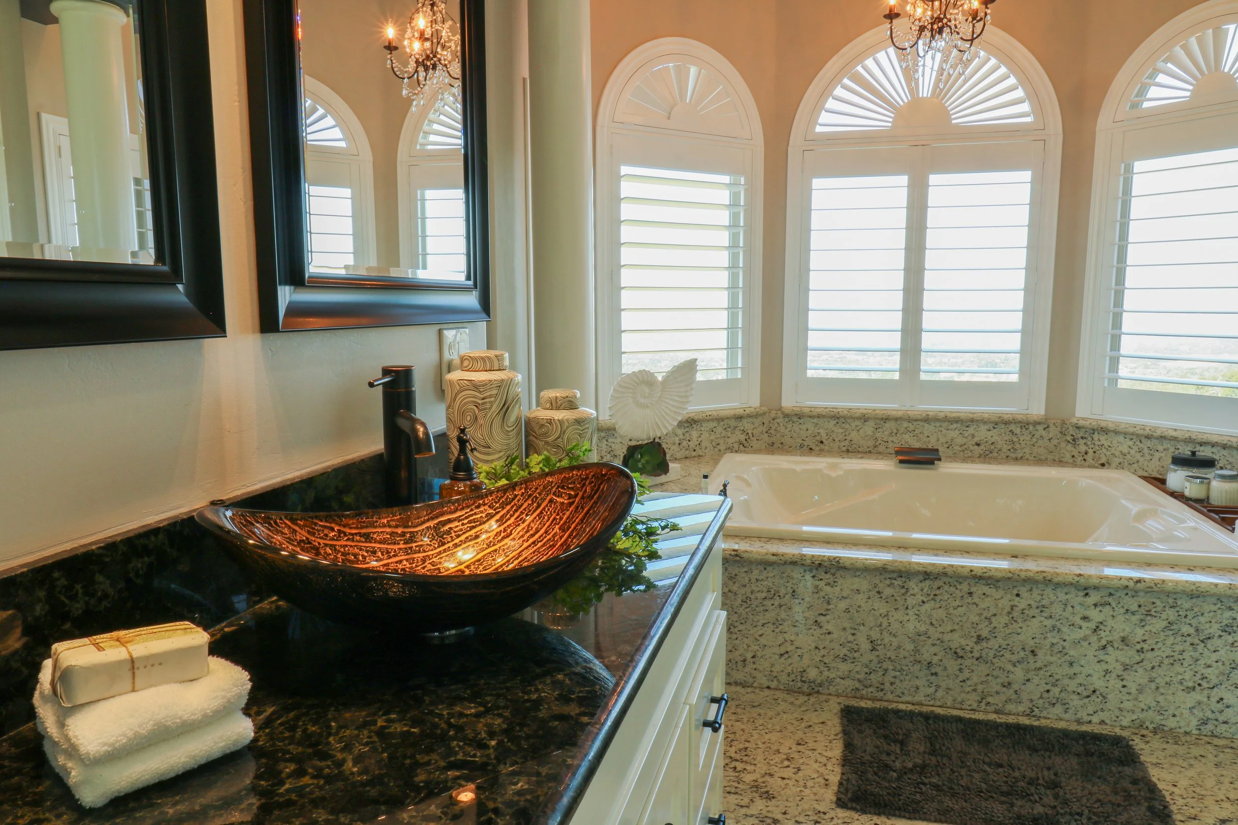 Bathroom interior featuring a granite countertop with a decorative vessel sink, a countertop soap dispenser, a rolled towel, and a bar of soap. Behind, there are three large arched windows with white shutters, a corner bathtub, and two black-framed m