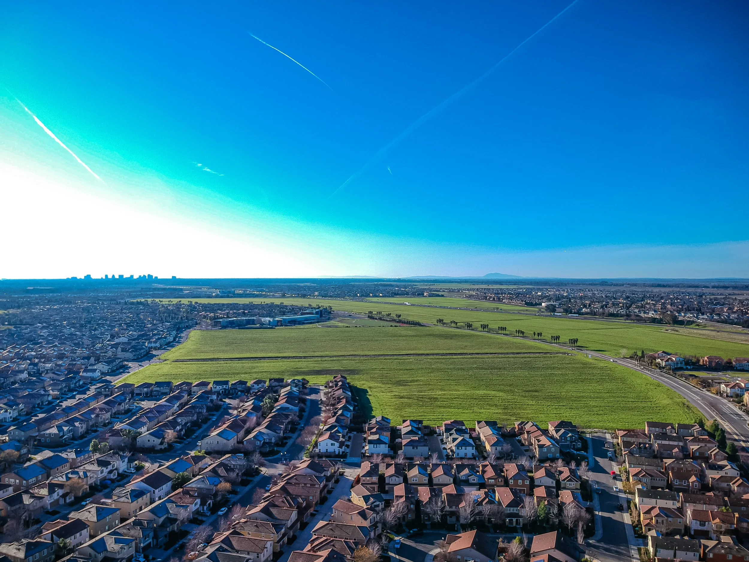 Aerial view of a suburban neighborhood adjacent to green fields under a clear blue sky with a few contrails.