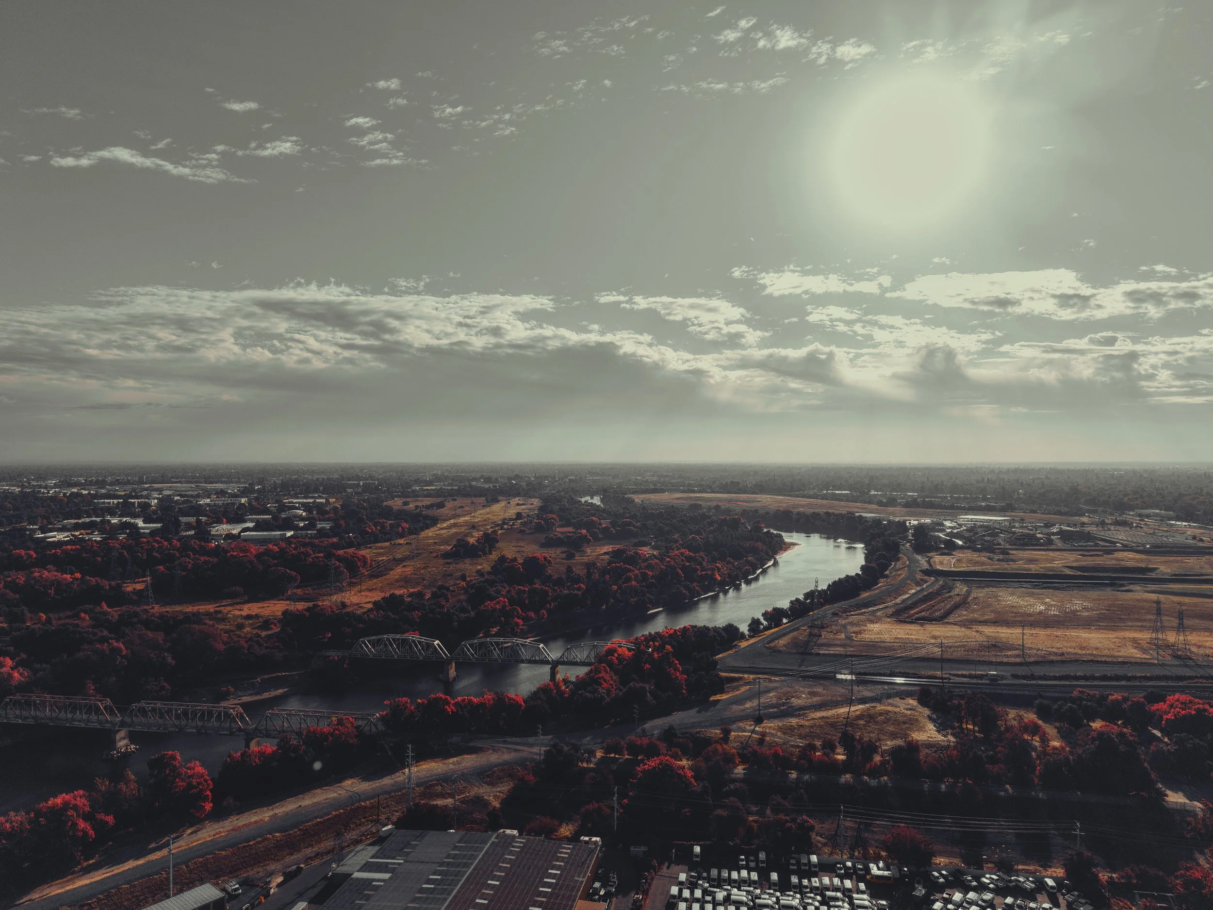 Aerial view of a river winding through a landscape with trees and fields, under a partly cloudy sky with the sun shining.