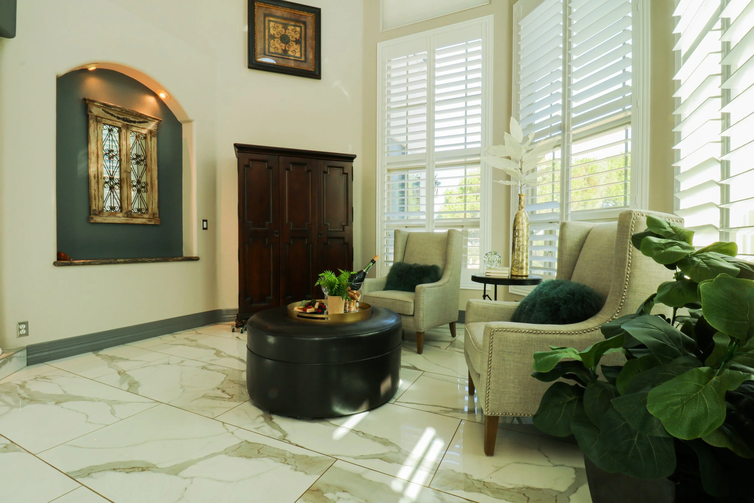 Living room with two beige armchairs, a large window with white shutters, a dark wooden wardrobe, green plants, and a black round ottoman with a tray of wine and snacks.