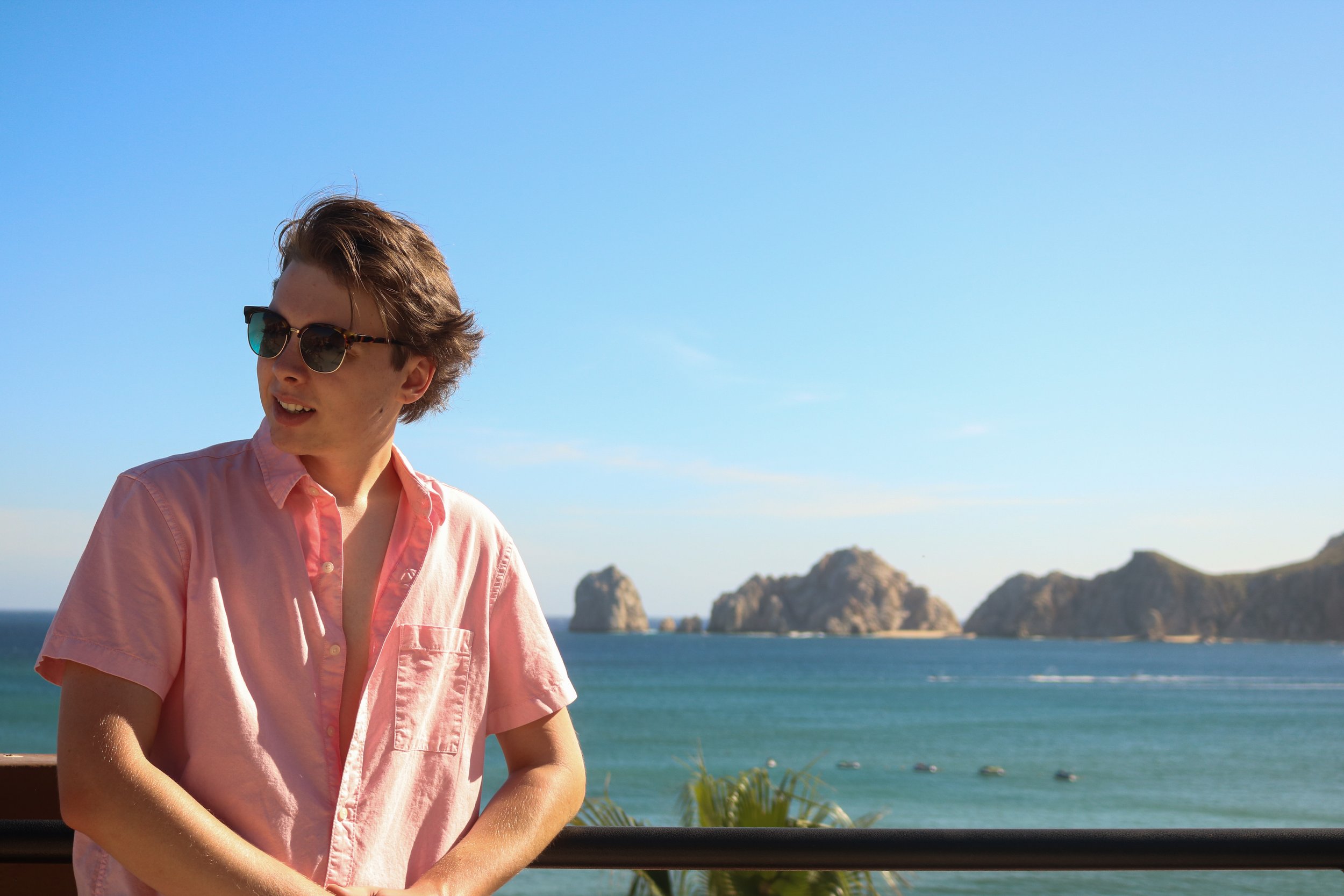 Young man wearing sunglasses and a pink short-sleeve shirt standing outdoors with ocean and rocky islands in the background on a sunny day.