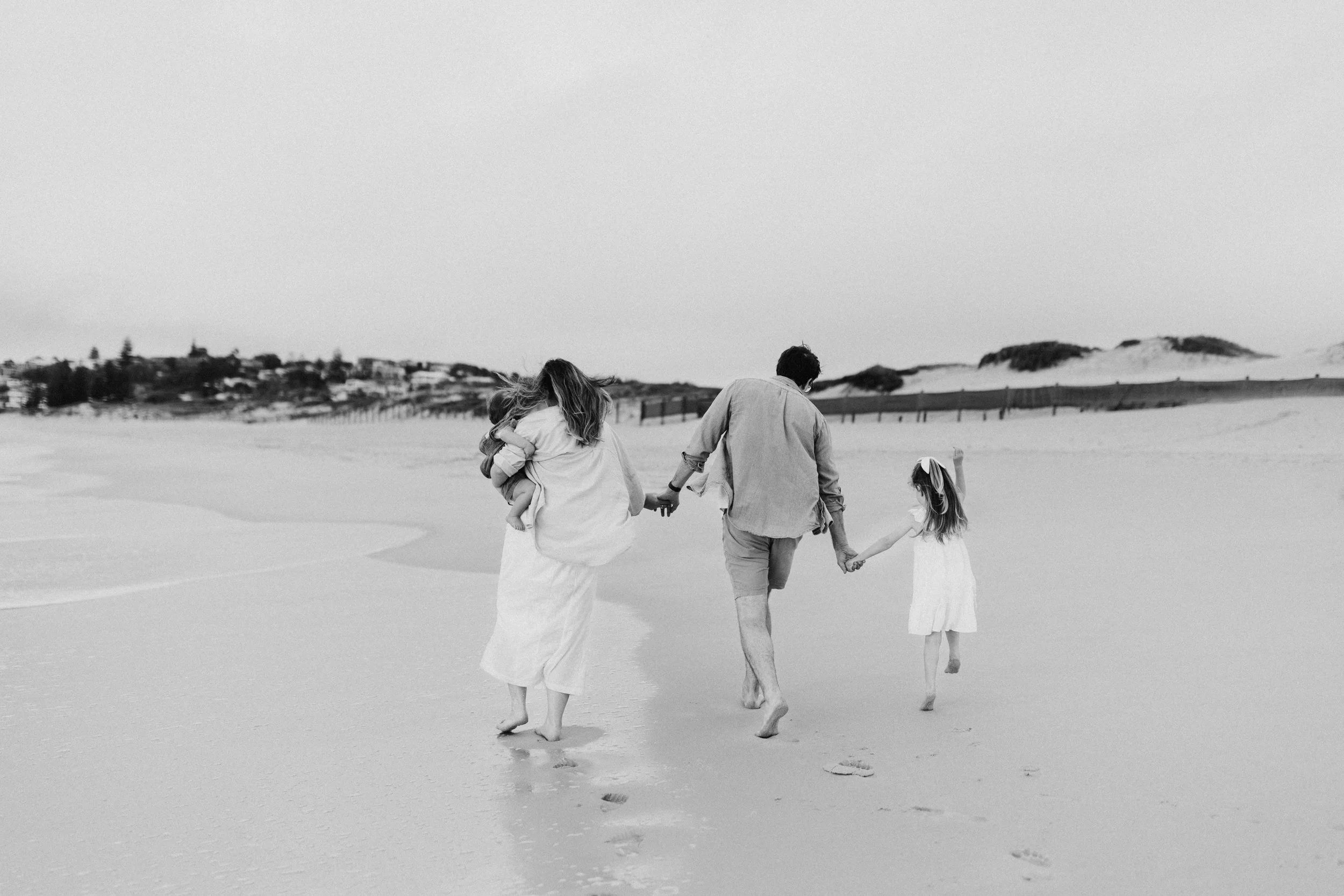 A family of four walking hand in hand on a sandy beach, with dunes and houses in the background, in a black-and-white photograph.