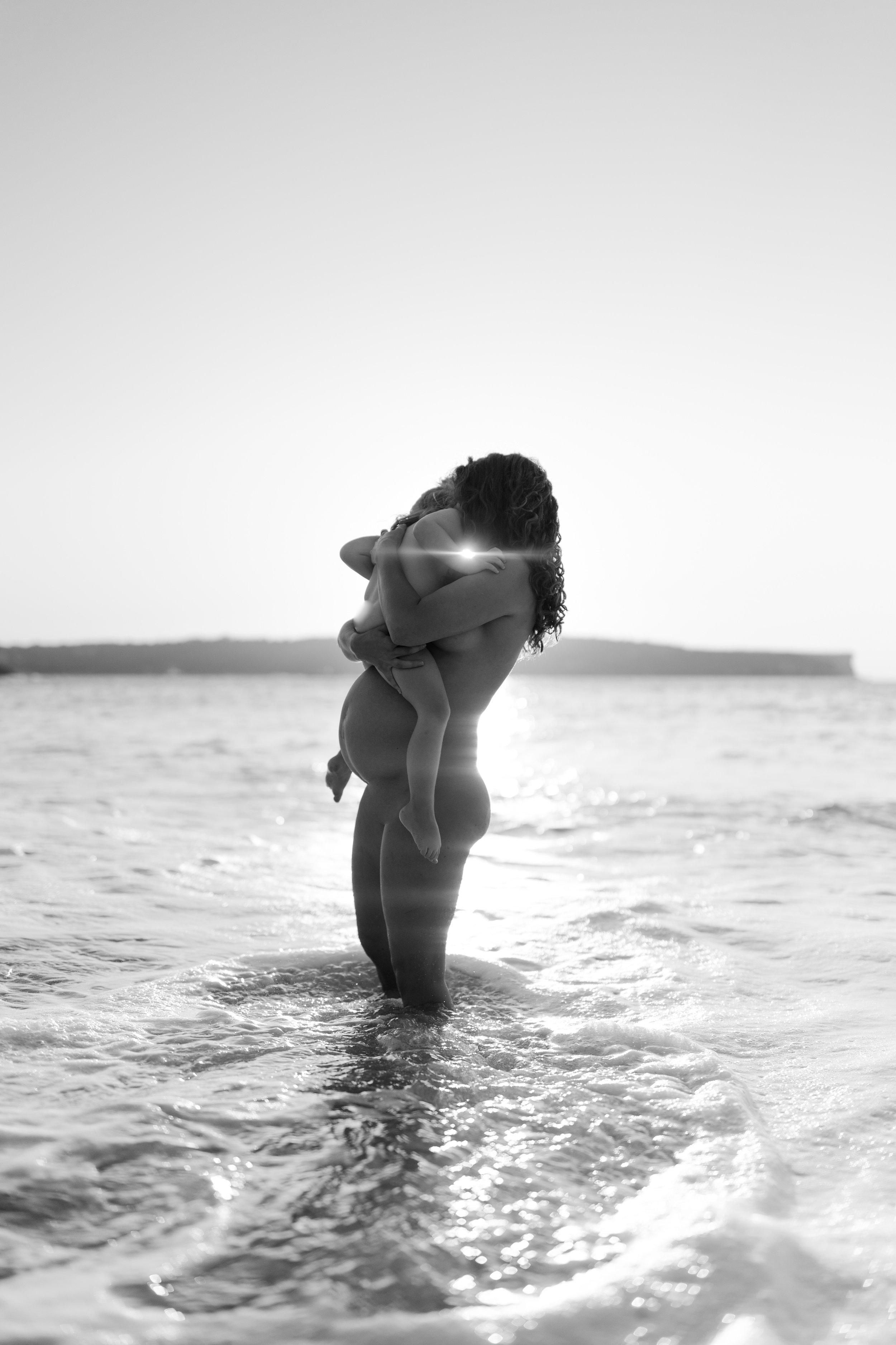 A person standing in the ocean holding a child, both naked, with the sun setting in the background, creating a silhouette effect in black and white.