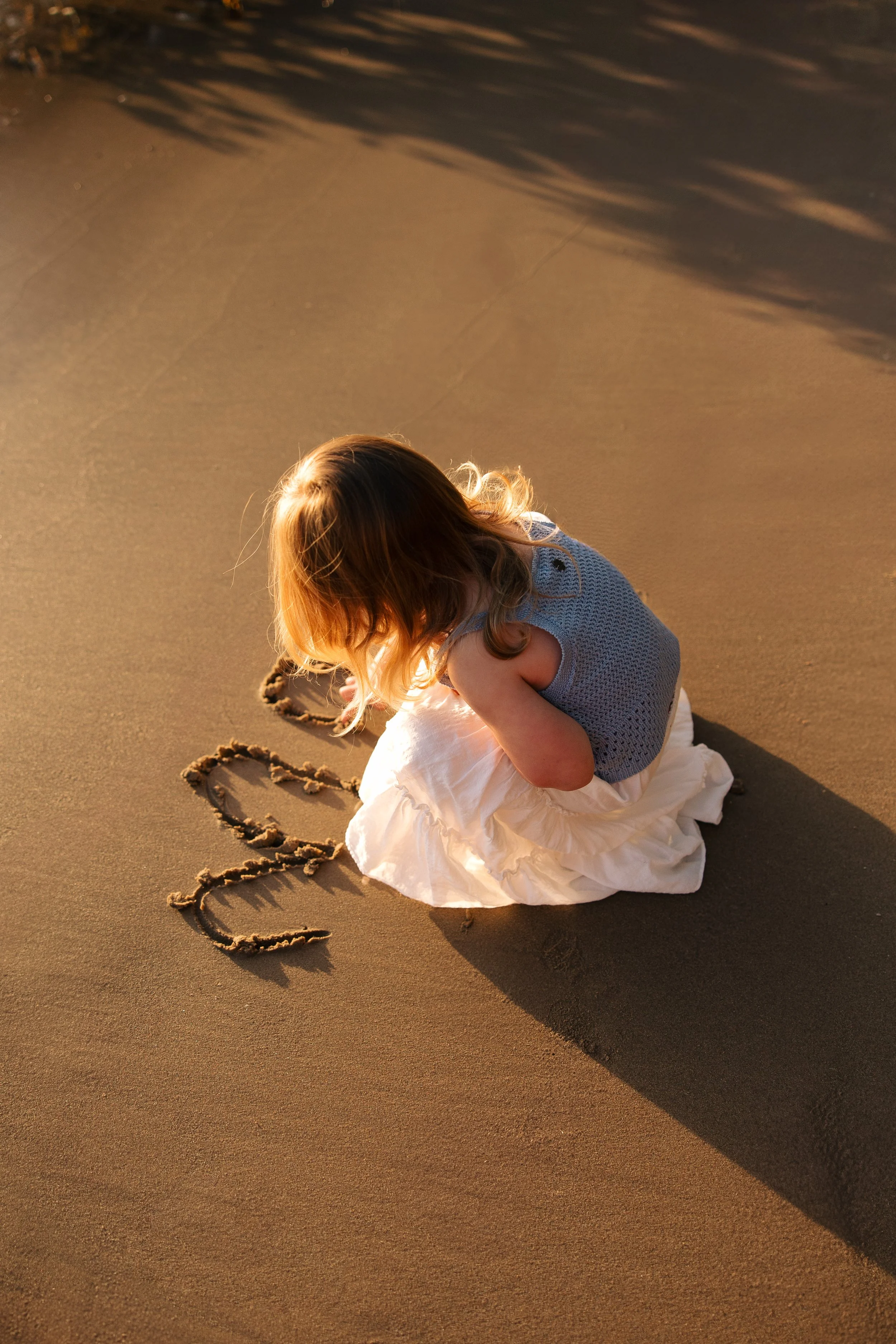 A young girl with red hair, wearing a blue sleeveless top and white skirt, sitting on the ground with her head bowed, writing the name 'Lola' in the sand.