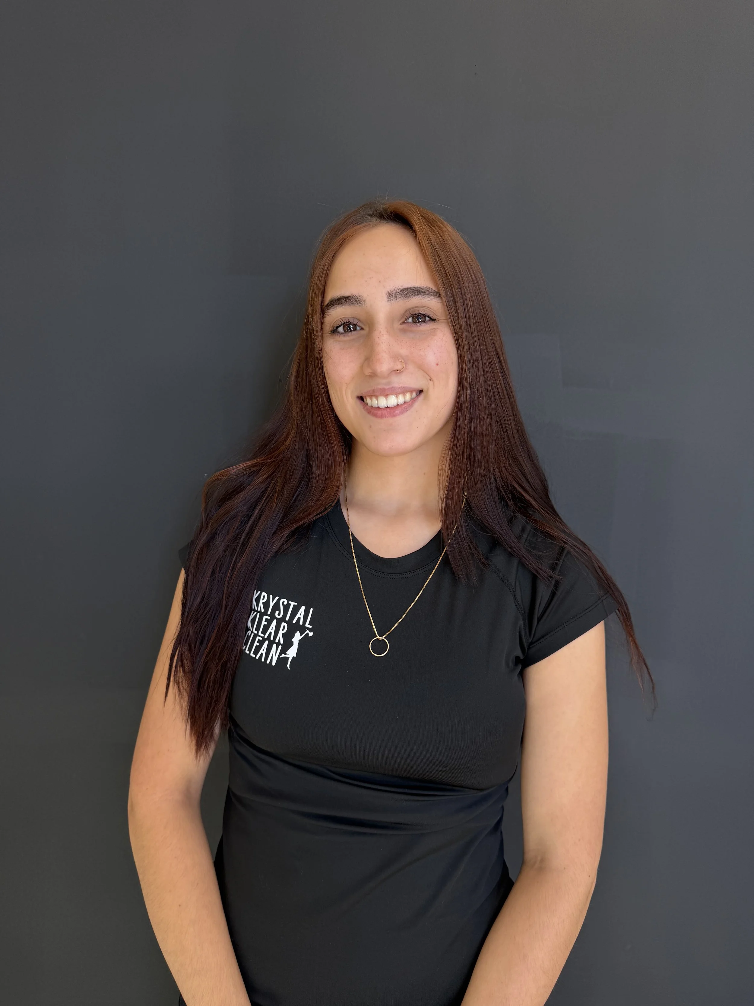 A young woman with brown hair in loose waves, wearing earrings and a black zip-up athletic jacket, standing against a dark gray background and smiling at the camera.