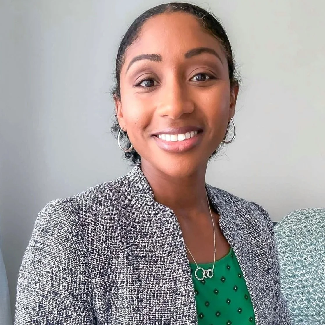 A woman with dark curly hair pulled back, wearing a gray patterned blazer over a green top with small black dots, silver hoop earrings, and a silver necklace, smiling at the camera against a plain light background.