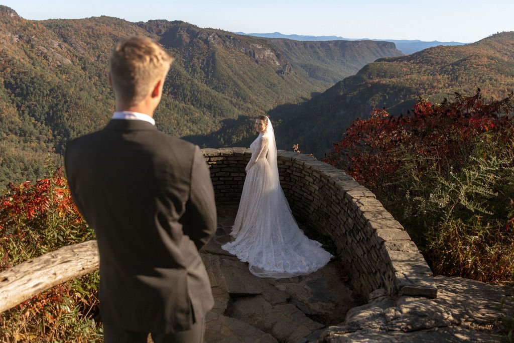 A couple in wedding attire stands on a stone viewing platform overlooking a mountain valley with autumn foliage.