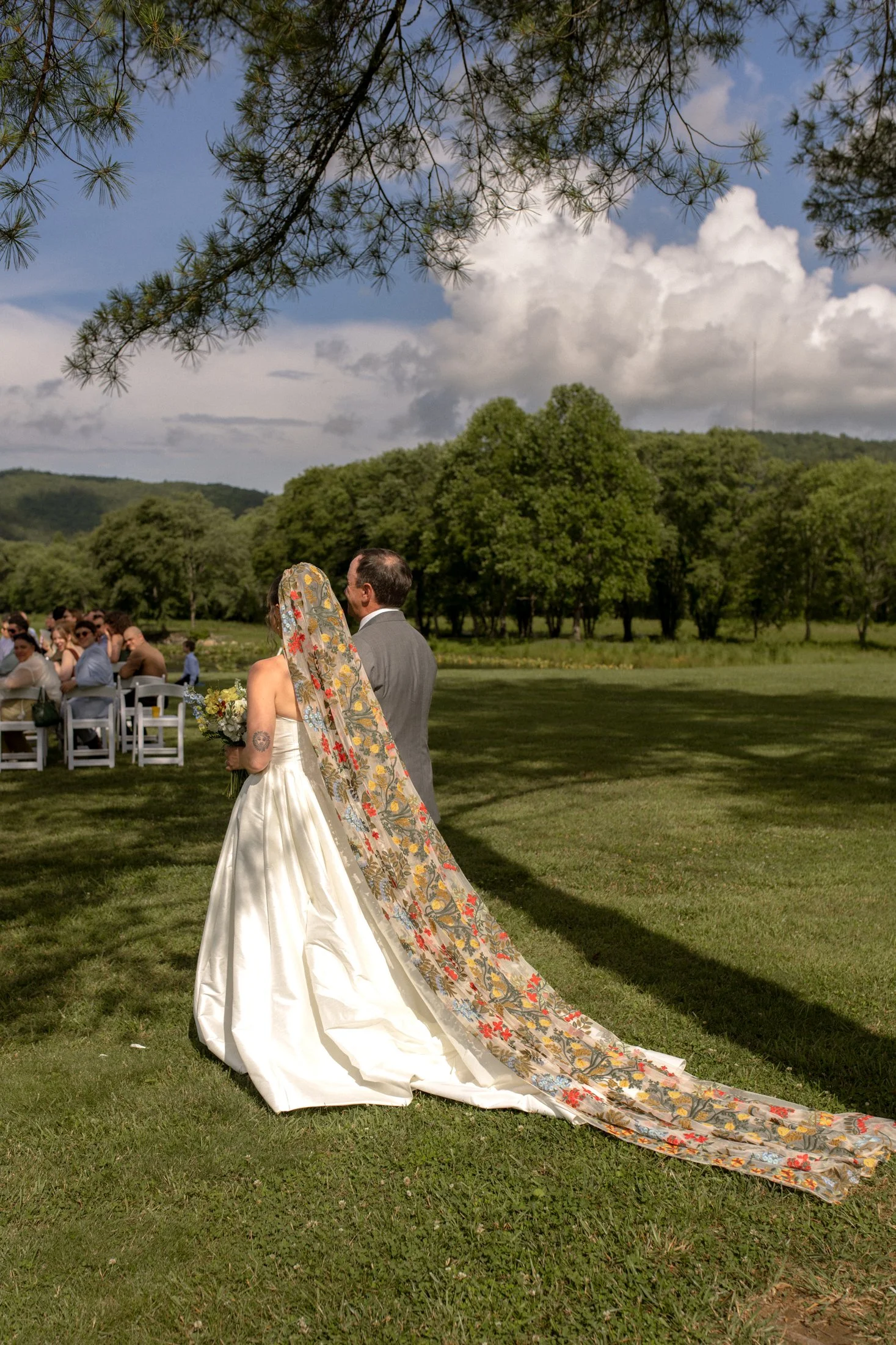 A bride and groom exchanging vows outdoors during a wedding ceremony on a grassy field with trees and a blue sky in the background.