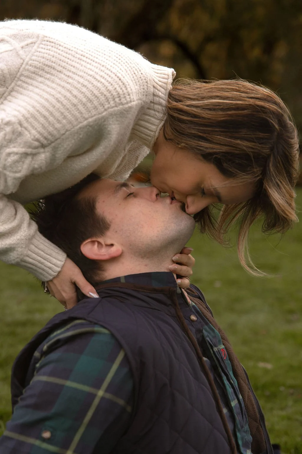 A woman leaning over and kissing a man on the lips outdoors during autumn, with green grass and blurred trees in the background.