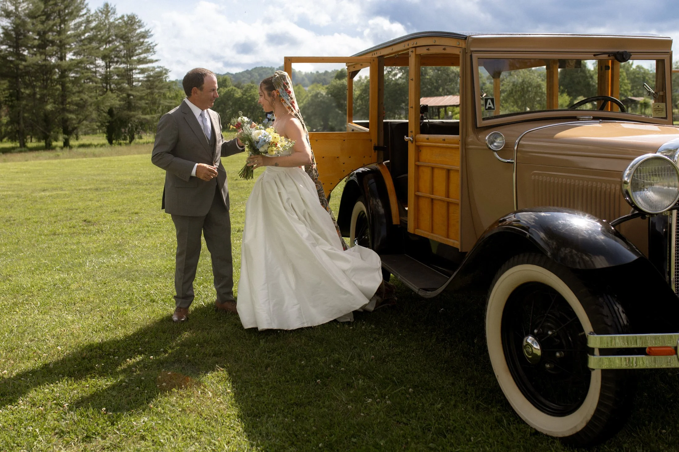 A bride in a white wedding dress holding a bouquet talking to a man in a suit next to a vintage wooden and black automobile in a grassy field with trees and hills in the background.