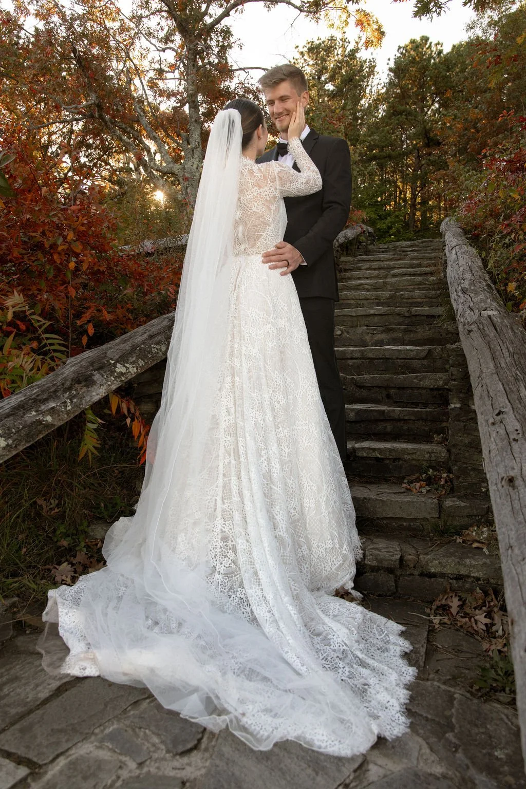 A bride and groom standing on a stone staircase outdoors, sharing a romantic moment at sunset surrounded by autumn foliage. The bride wears a white lace wedding gown with a long veil, and the groom is dressed in a black tuxedo. The couple looks happy