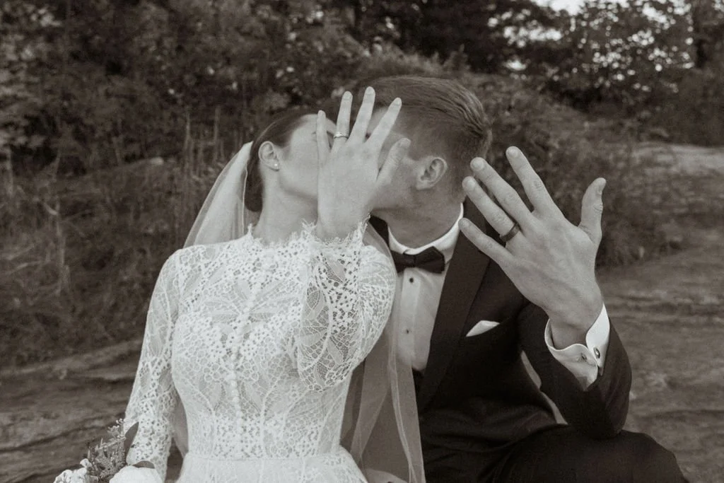A bride and groom are kissing outdoors, both showing their wedding rings while covering their faces with their hands in front of the camera.