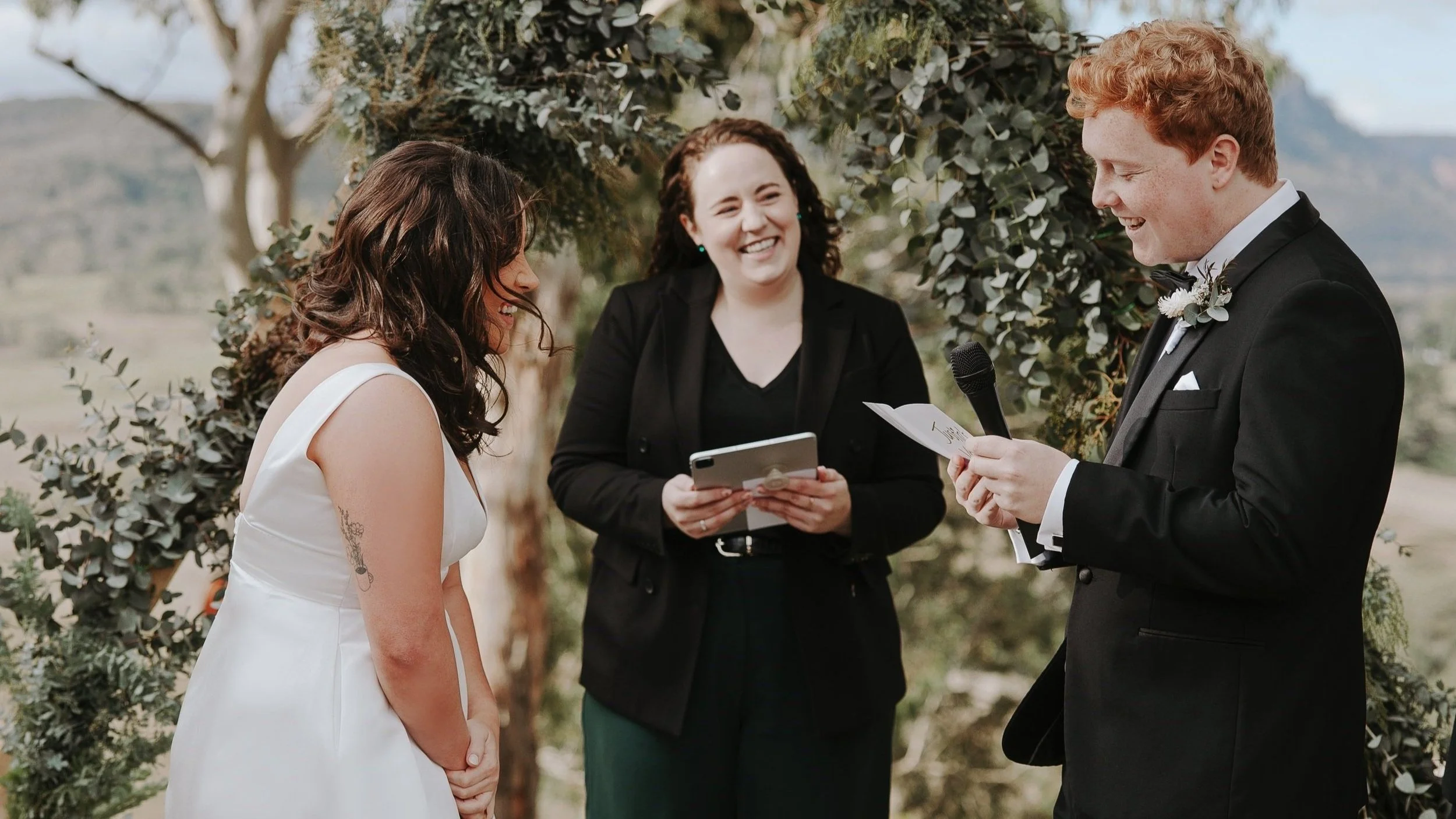 A bride in a white gown with a tattoo on her arm, and a groom in a black tuxedo, are laughing during their vows with Elle standing beside them. They are under a eucalyptus arch, with the vineyards of Dryridge Estate in the Megalong Valley behind them