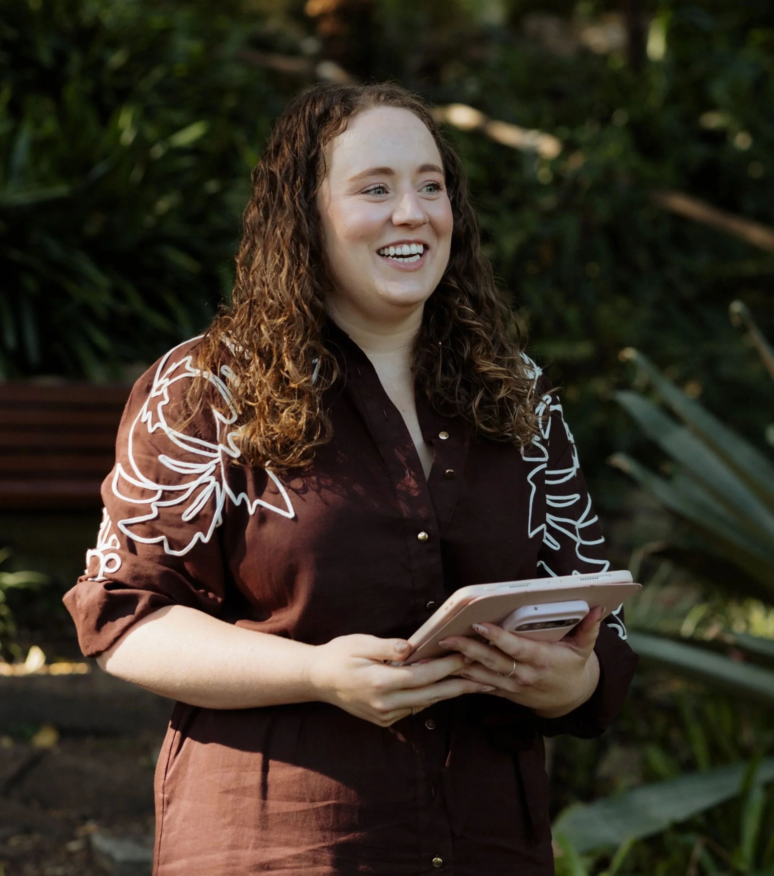 Elle the celebrant is wearing a brown dress with white embroidery and her long curly brown hair is out and standing in a tropical garden holding her iPad and smiling during a couple's elopement ceremony in Sydney
